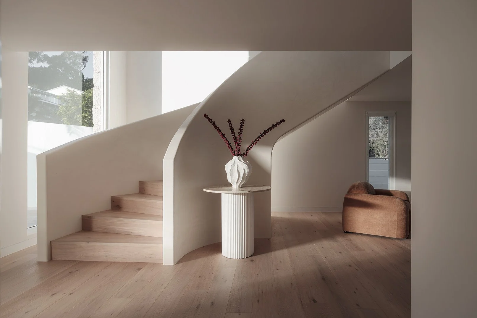 Interior of a modern home featuring a curved staircase with light wood steps and white walls, a white pedestal with a white sculptural vase holding purple flowers, and a brown armchair against the wall near a window.