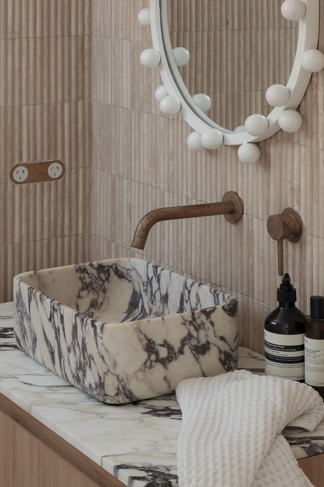 Bathroom sink area with a marble vessel sink, bronze faucet, mirror with white round bulb lights, beige textured wall, black soap dispenser, and a folded white towel.