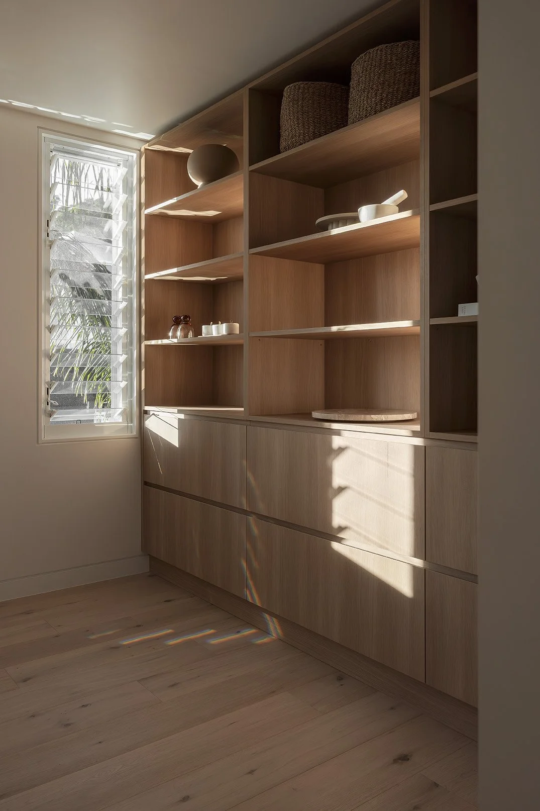 Corner of a room with a wooden shelving unit and drawers, sunlight casting shadow lines, a window with louvered blinds showing plants outside.