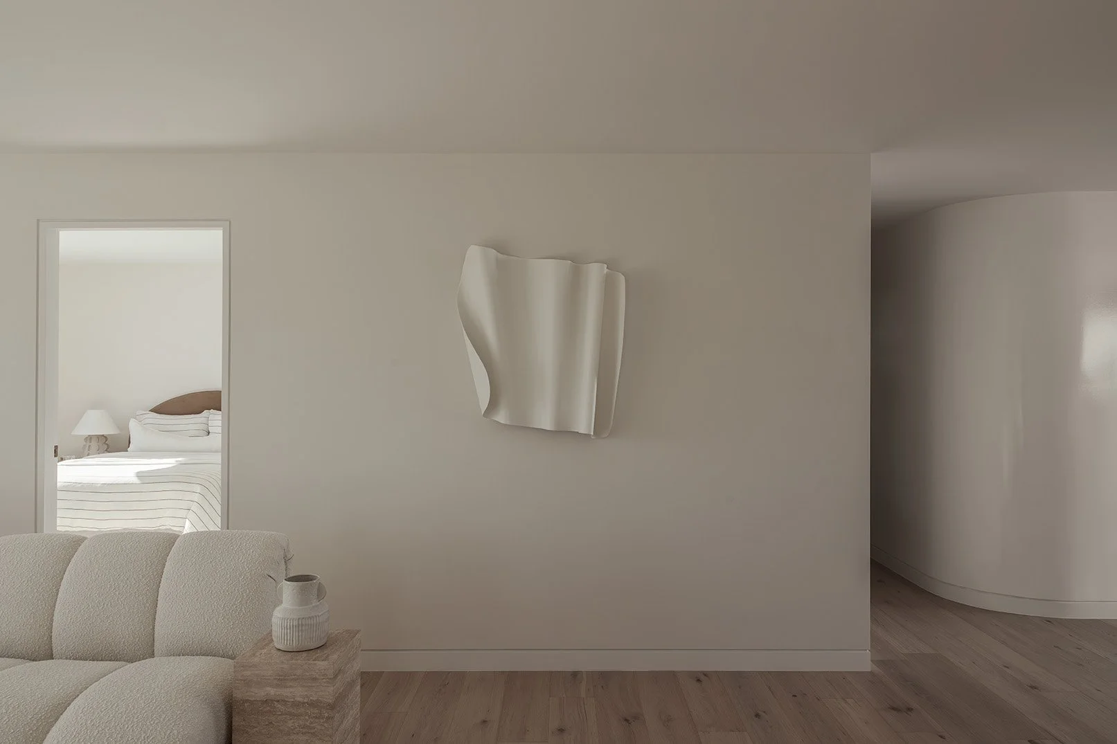 Minimalist living room with a cream-colored sofa, a small wooden side table with a ceramic vase, a wall-mounted abstract sculpture, and a doorway leading to a bedroom with a bed and a nightstand with a lamp.