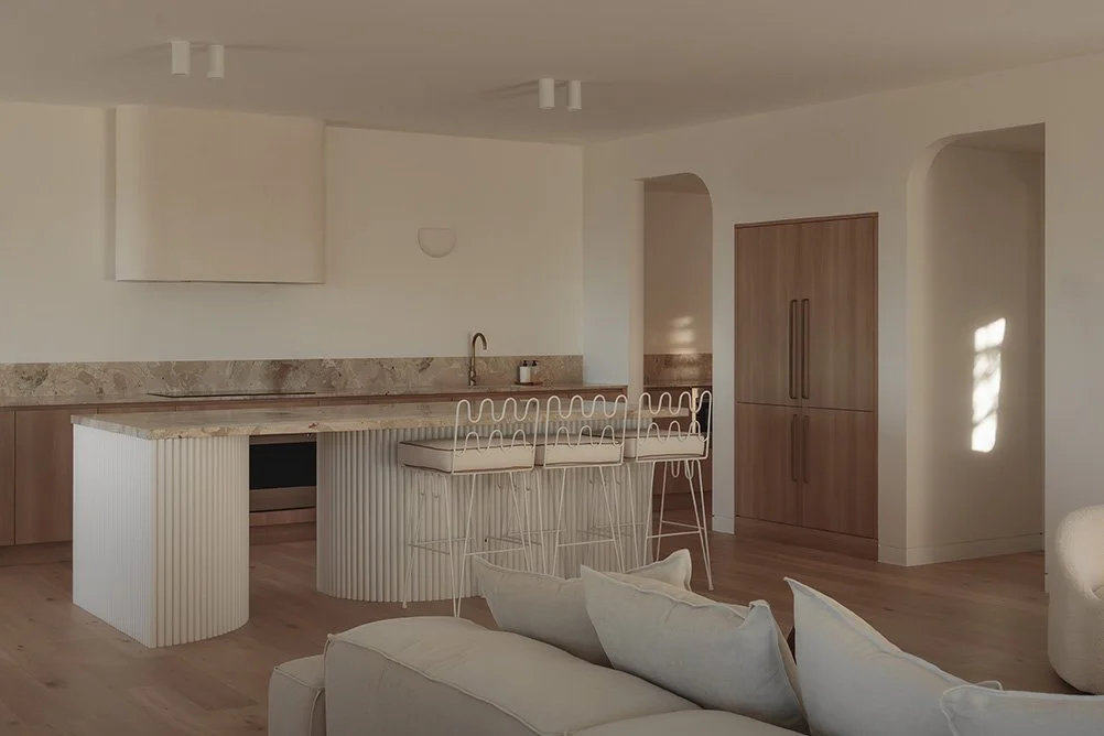 Modern minimalist kitchen with beige walls, wooden cabinetry, and a marble backsplash. White bar stools are at the kitchen island, and a section of a white sofa with pillows is in the foreground.