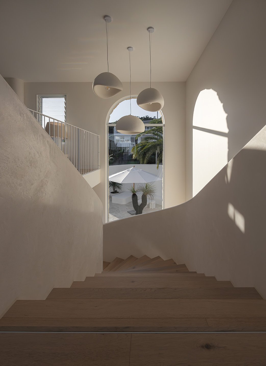 Interior view of a staircase landing with a large arched window, white walls, wooden stairs, and modern hanging pendant lights. Outside, there is a patio with an umbrella and a cactus, palm trees, and neighboring houses.