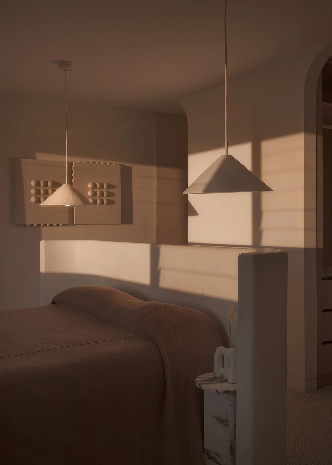 A bedroom with a bed, a small side table with a kitchen kettle, two hanging pendant lights, and sunlight casting shadows on the wall.