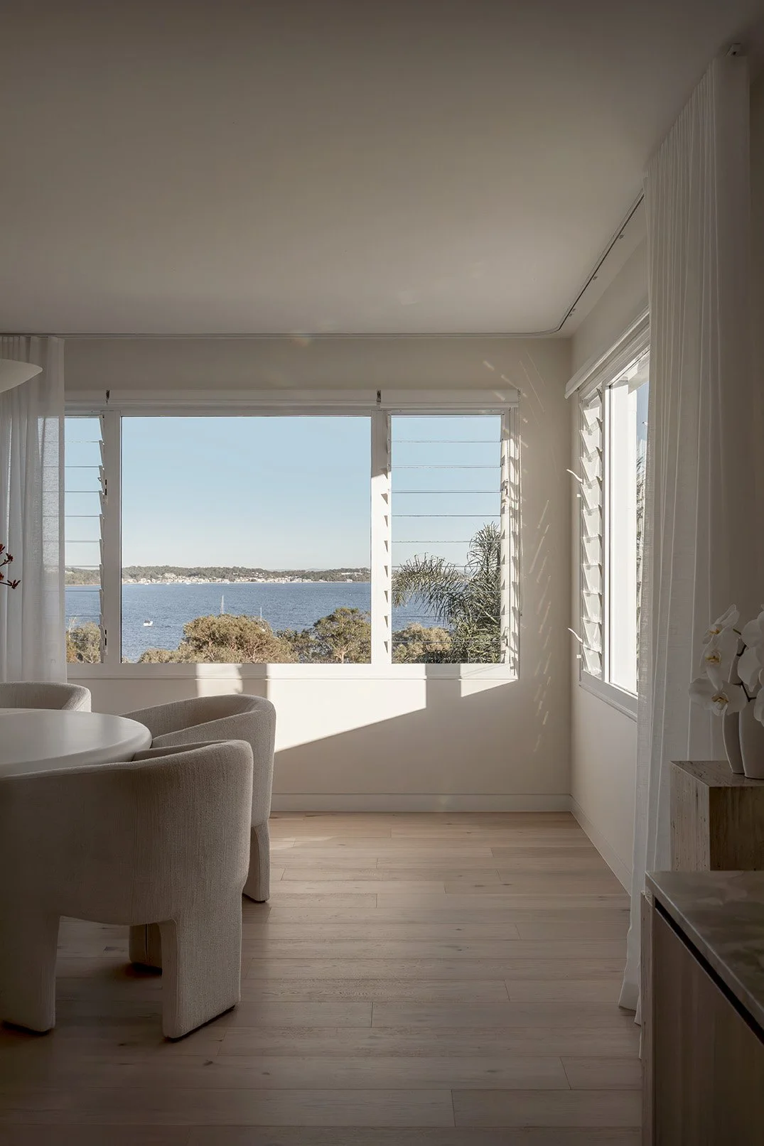Interior of a room with large windows overlooking a body of water and trees, sunlight casting shadows on the floor, white curtains, wooden floor, and part of a white chair and a vase with white flowers visible.