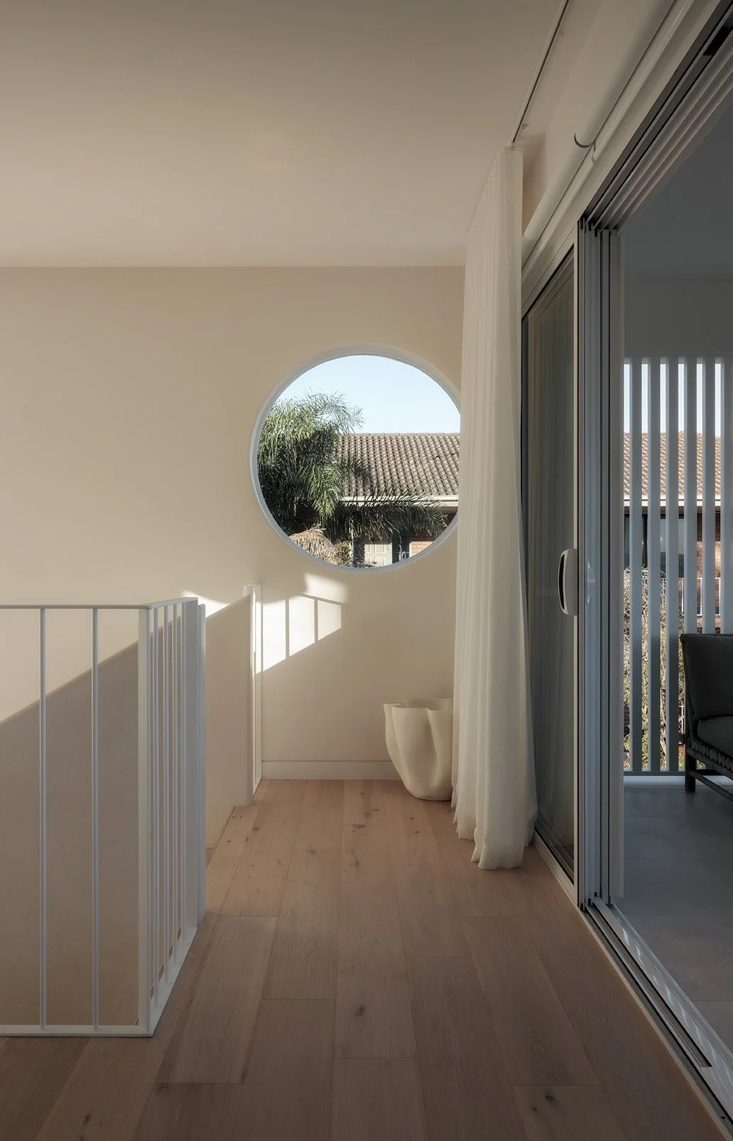 A modern indoor balcony area with a round window, white curtains, a plant, and sliding glass doors leading to an outdoor patio.
