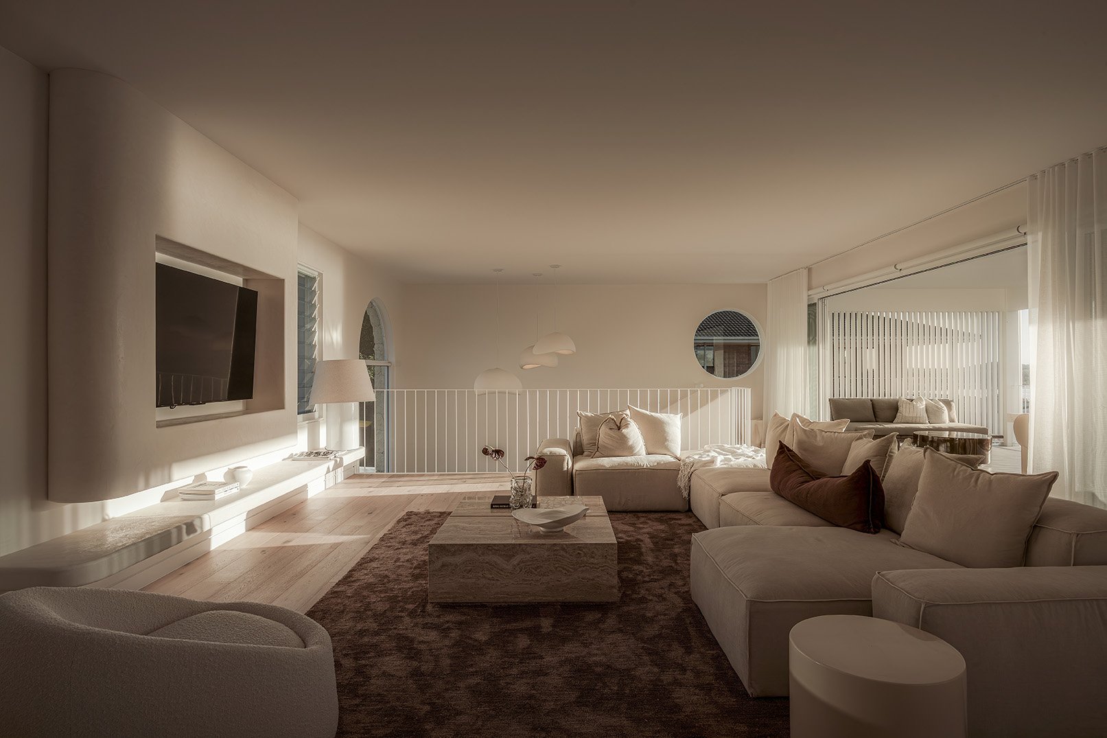 Bright living room with white walls, beige sectional sofa, round mirror, TV on the wall, large windows with white curtains, wooden coffee table, and a brown rug.