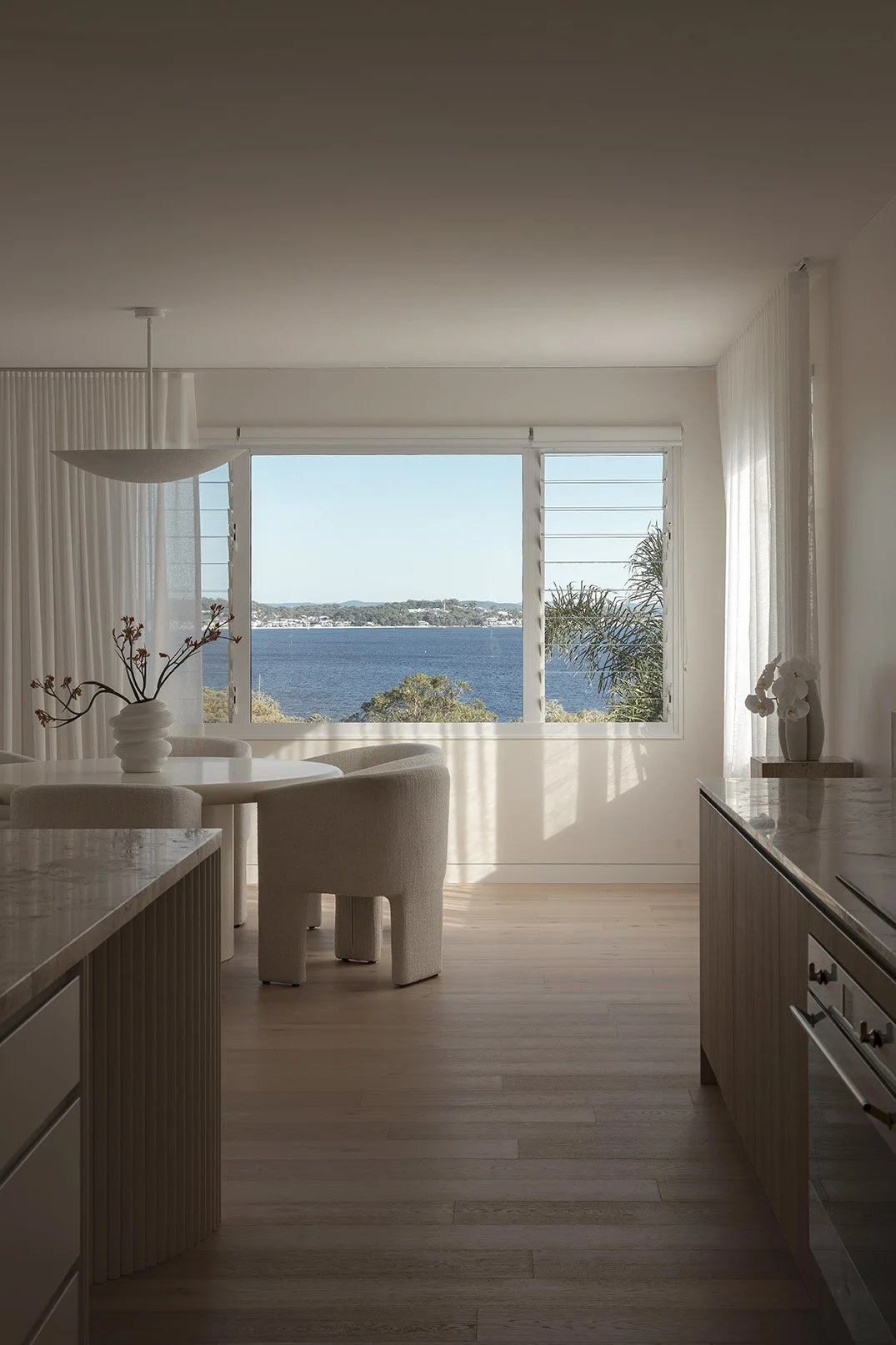 Interior view of a bright, minimalist room with a window overlooking a body of water and greenery outside. The room has light-colored wooden flooring, white curtains, a round dining table with four chairs, and a sideboard with decorative vases and fl