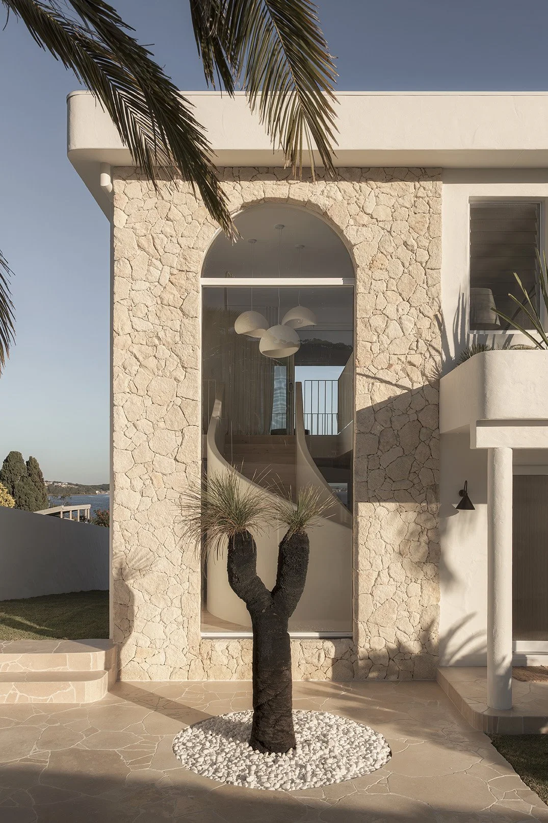 Front view of a modern, minimalist house with stone facade, large arched window, a decorative tree with black trunk, white rocks, and tropical palm leaves in the foreground.