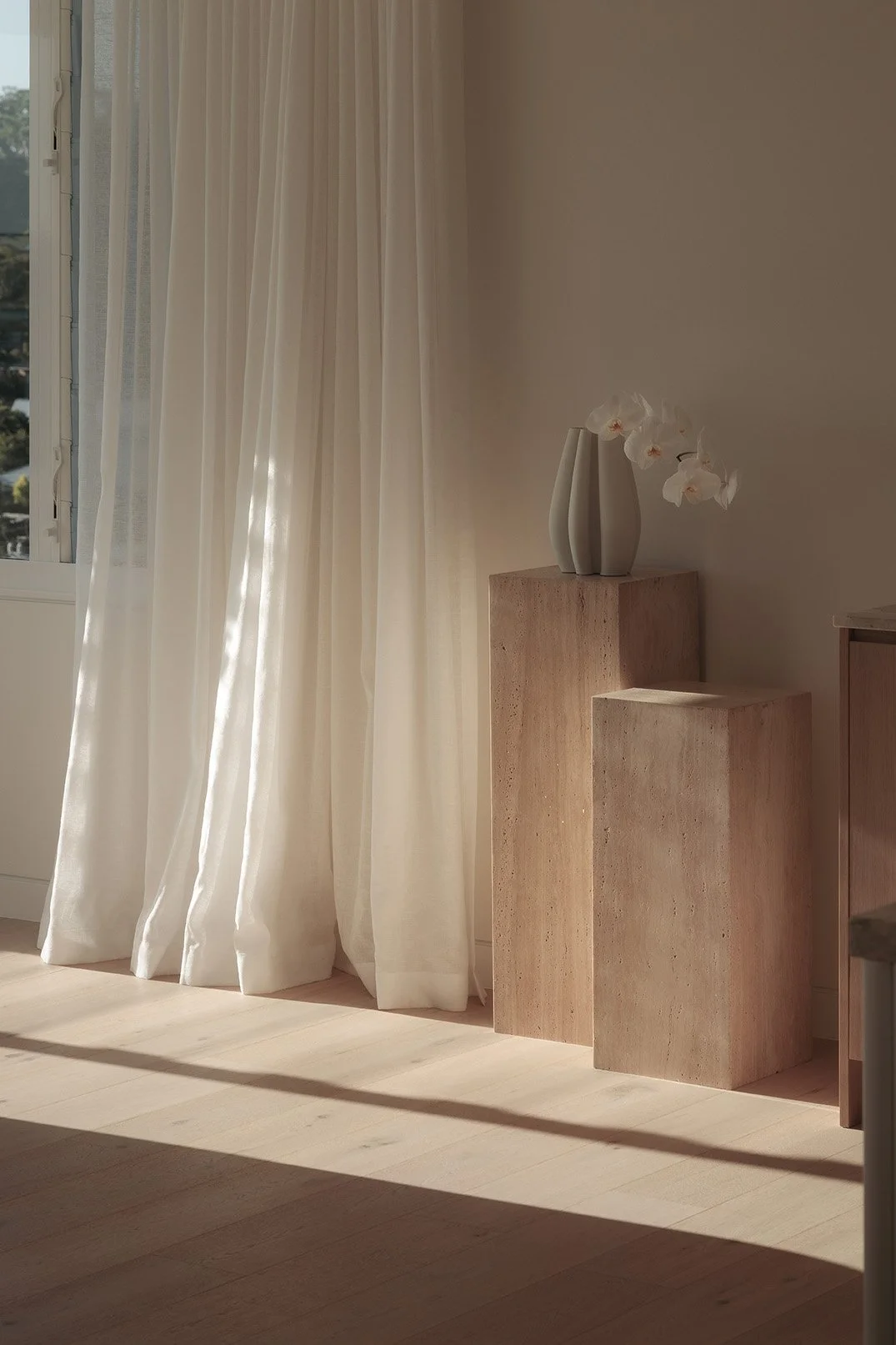Interior of a room with sunlight streaming through sheer white curtains, beige wooden floor, and two textured beige pedestals with a white orchid in a white vase on the taller pedestal.