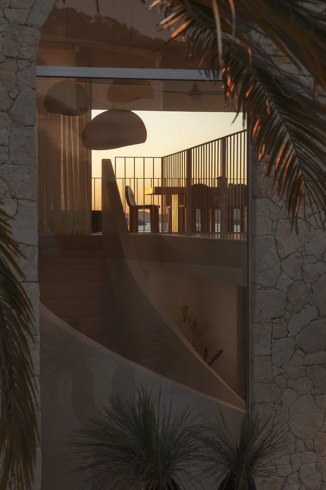 View of a balcony through a glass window with sunset in the background, including furniture like chairs and a table, and some interior lighting fixtures visible inside.