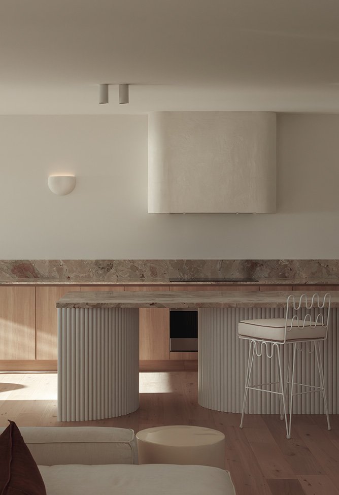 Modern interior kitchen with a curved wooden island, white wireframe chair, beige sofa, and minimalist wall lighting.