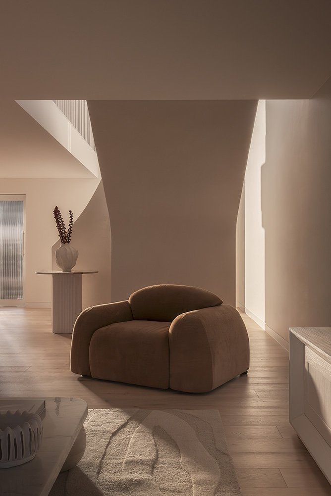 A cozy living room with a rounded brown armchair in front of a large light-filled window, with a beige rug and a side table with a vase of flowers in the background.
