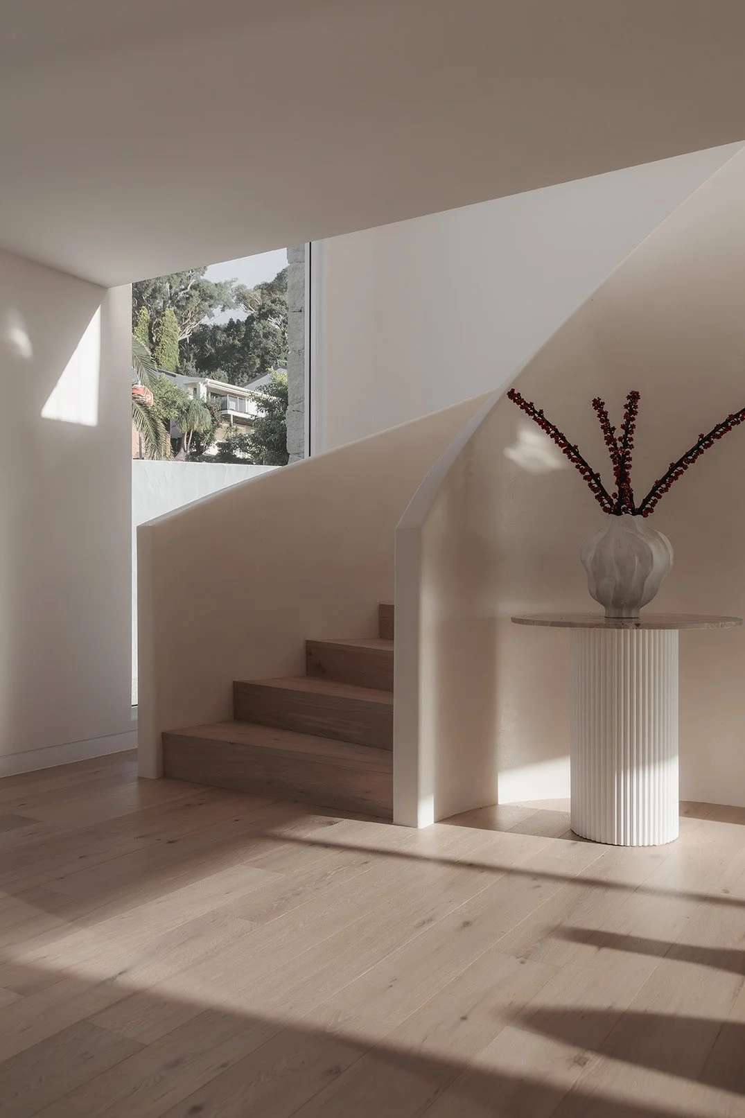 Sunlight streams into a minimalist interior with a wooden staircase, white walls, and a tall vase with dark red flowers on a textured pedestal table.