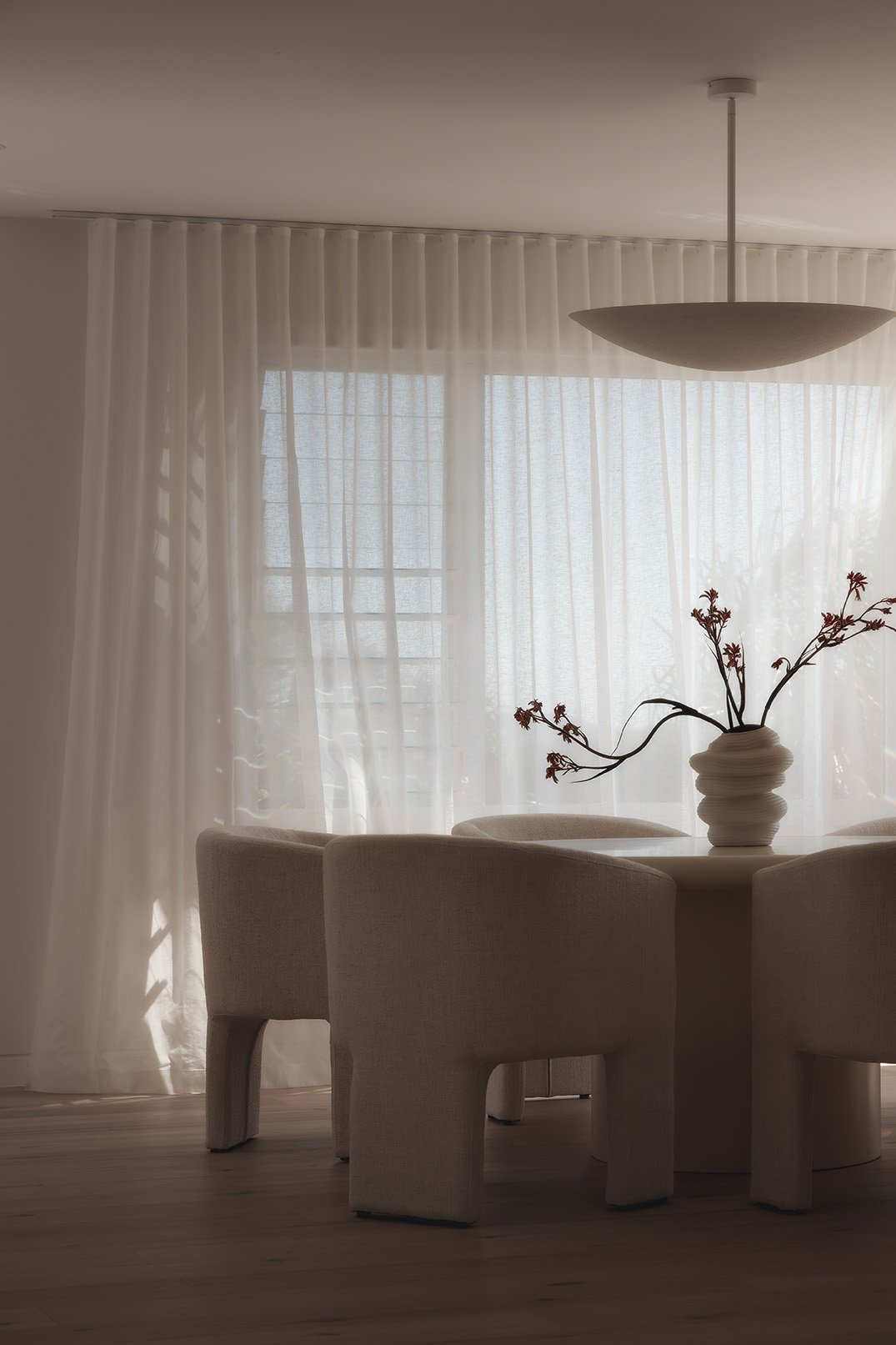 A dining area with beige upholstered chairs around a round table, a white vase with dried flowers, sheer white curtains, and a ceiling light fixture.