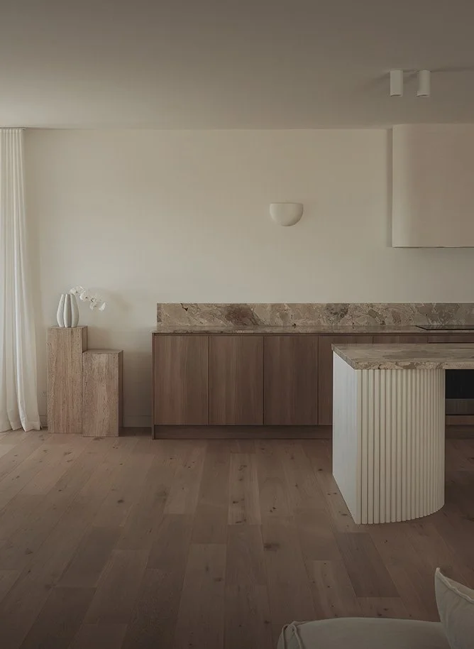 Minimalist kitchen with white walls, wooden cabinets, marble backsplash, and a small section of a white and wooden island.