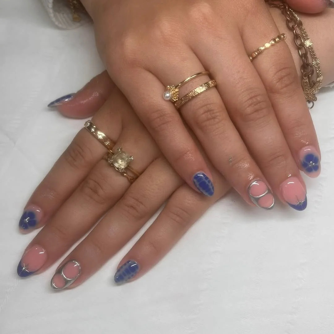 Close-up of two hands resting on a white surface, showing multiple gold rings and decorated nails. The nails are painted with blue and nude colors, featuring specific patterns, including grid-like designs and metallic accents. The rings include a pearl ring, a solitaire ring, and simple gold bands.