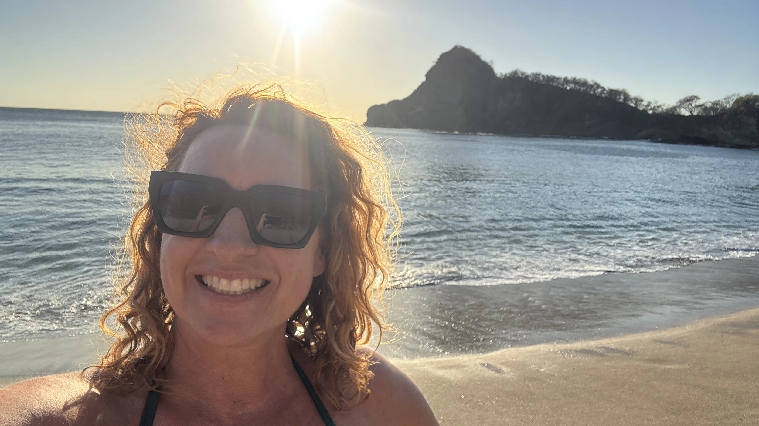 A woman with curly hair wearing sunglasses smiling at the beach during sunset, with the ocean and a rocky island in the background.