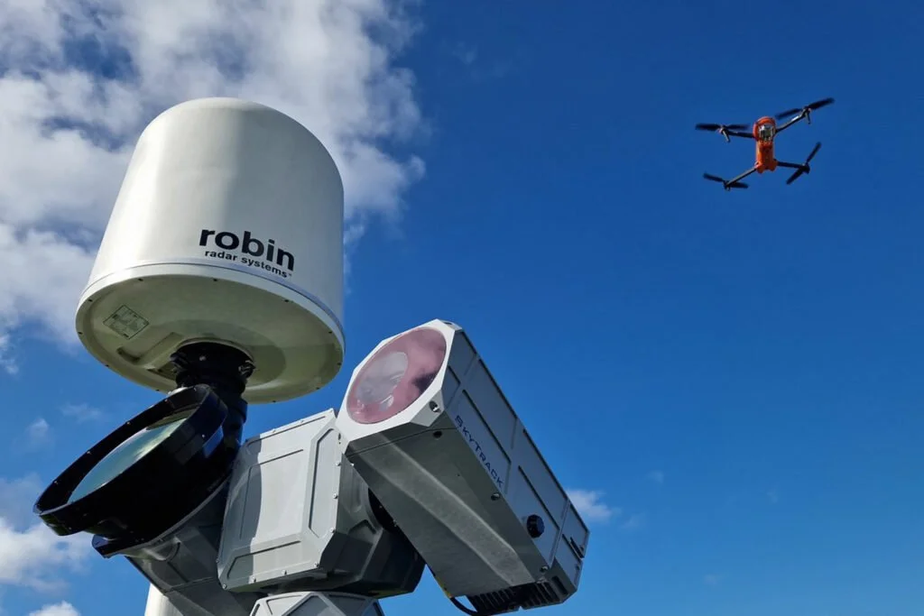 A radar system with a white dome labeled 'robin radar systems,' a camera or sensor device, and a drone flying in a partly cloudy sky.