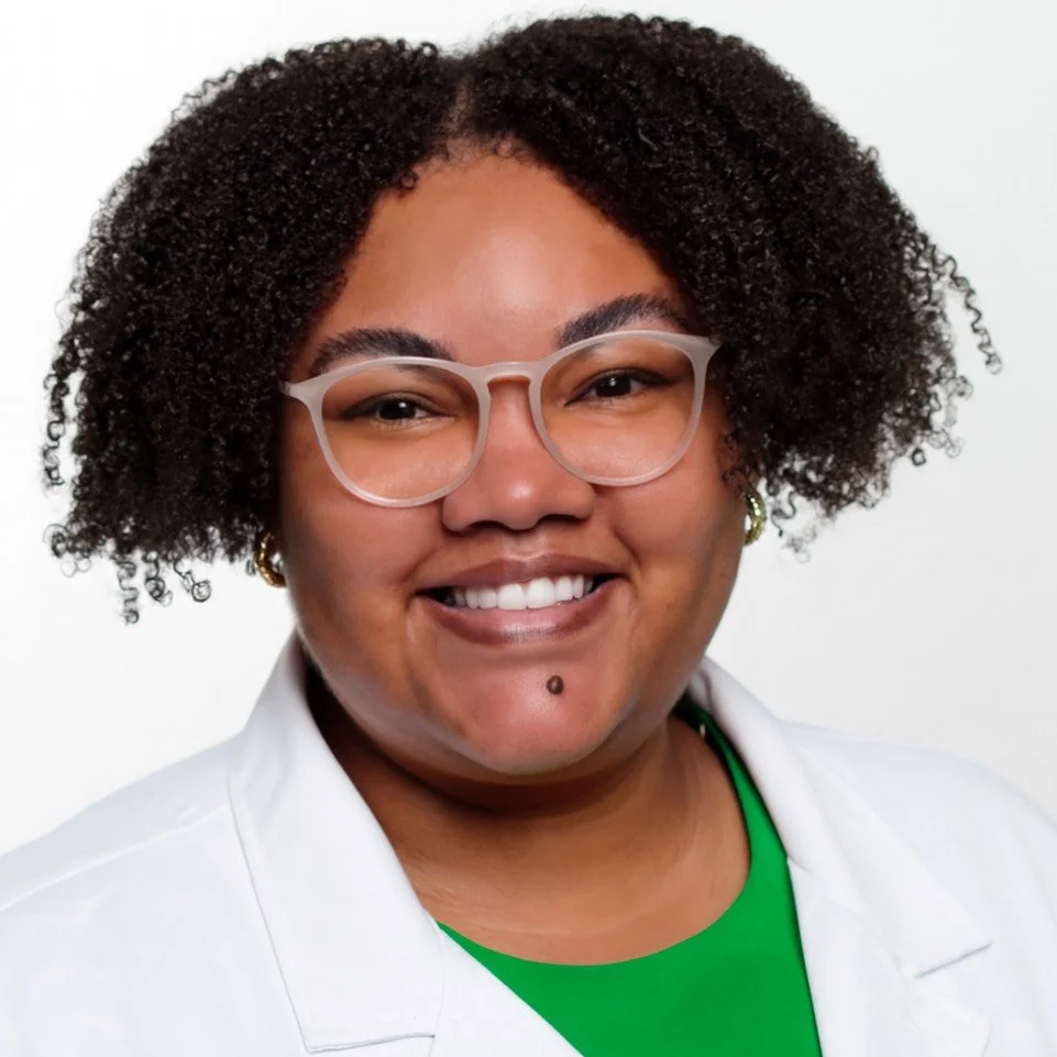 Close-up portrait of a woman with curly hair, glasses, and a bright smile, wearing a white coat and a green top.
