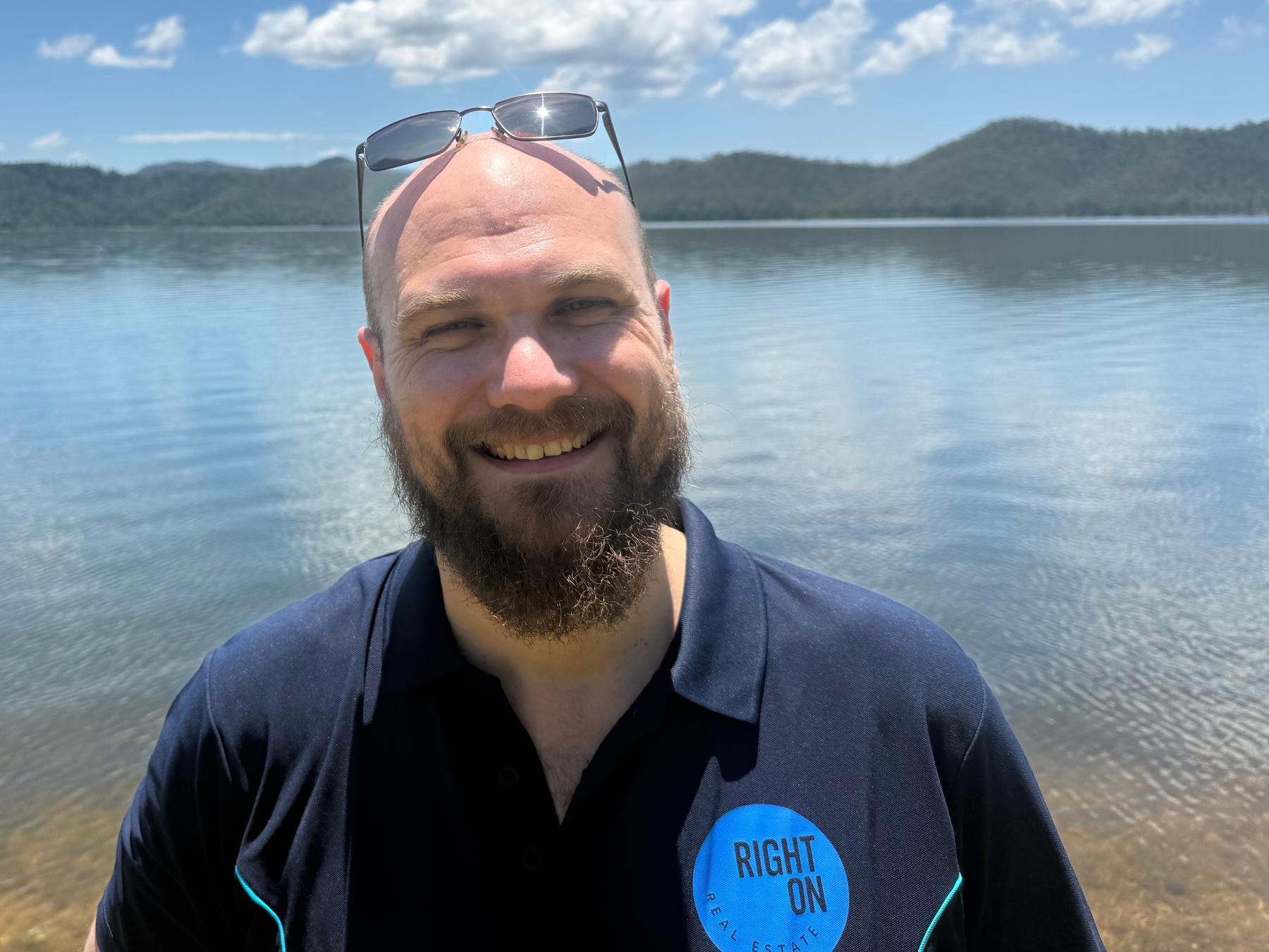 Man smiling near a lake with mountains in the background, wearing sunglasses on his head and a dark shirt with a 'Right On Real Estate' logo.