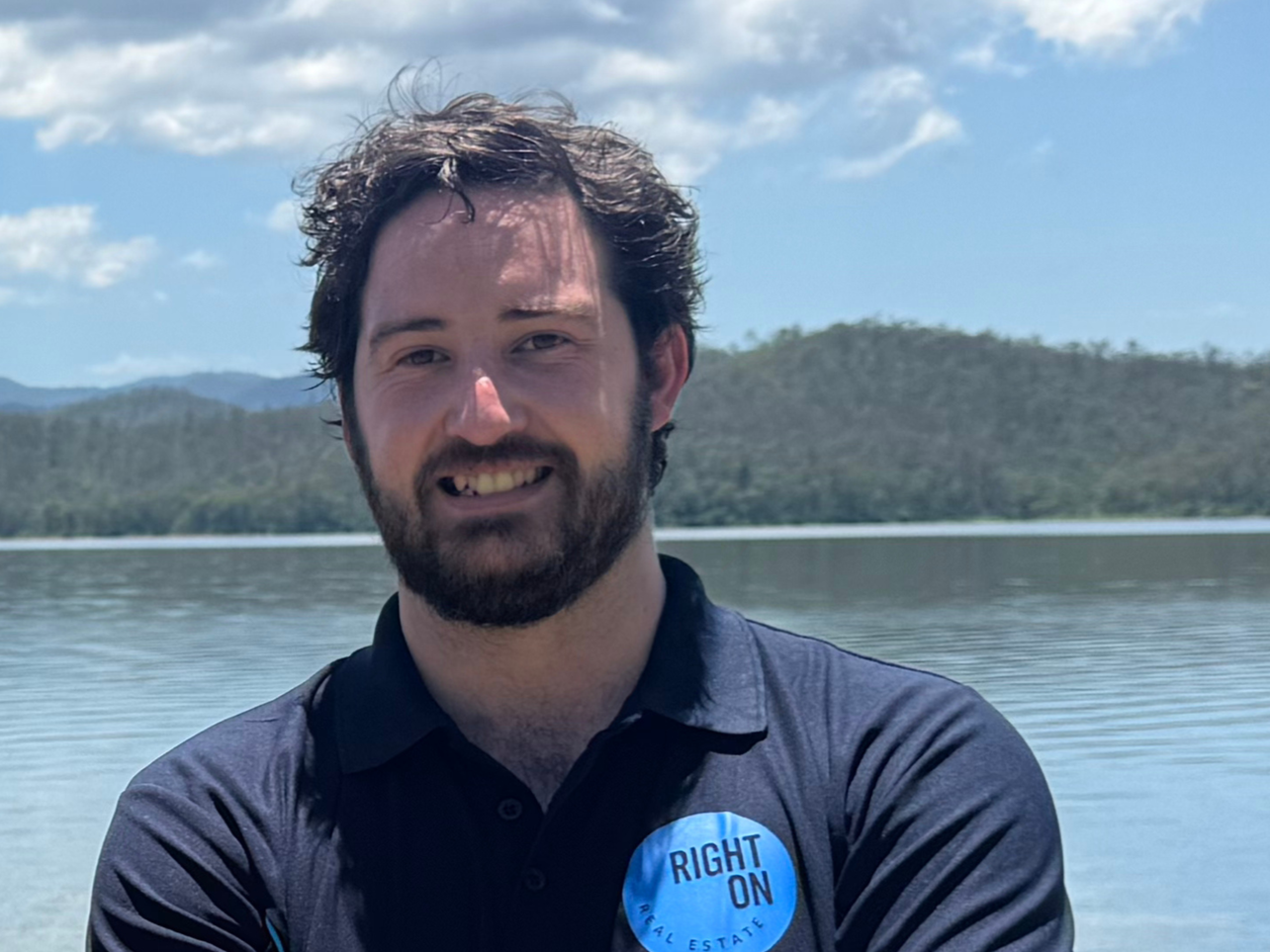 A man with dark curly hair and a beard, smiling, standing near a body of water with hills and a partly cloudy sky in the background, wearing a black shirt with a blue badge that says 'RIGHT ON REAL ESTATE.'