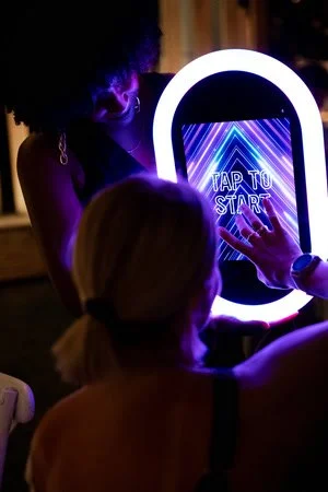 People playing an arcade game with a circular screen and a neon 'Tap to Start' message, illuminated by vibrant purple and blue lights.