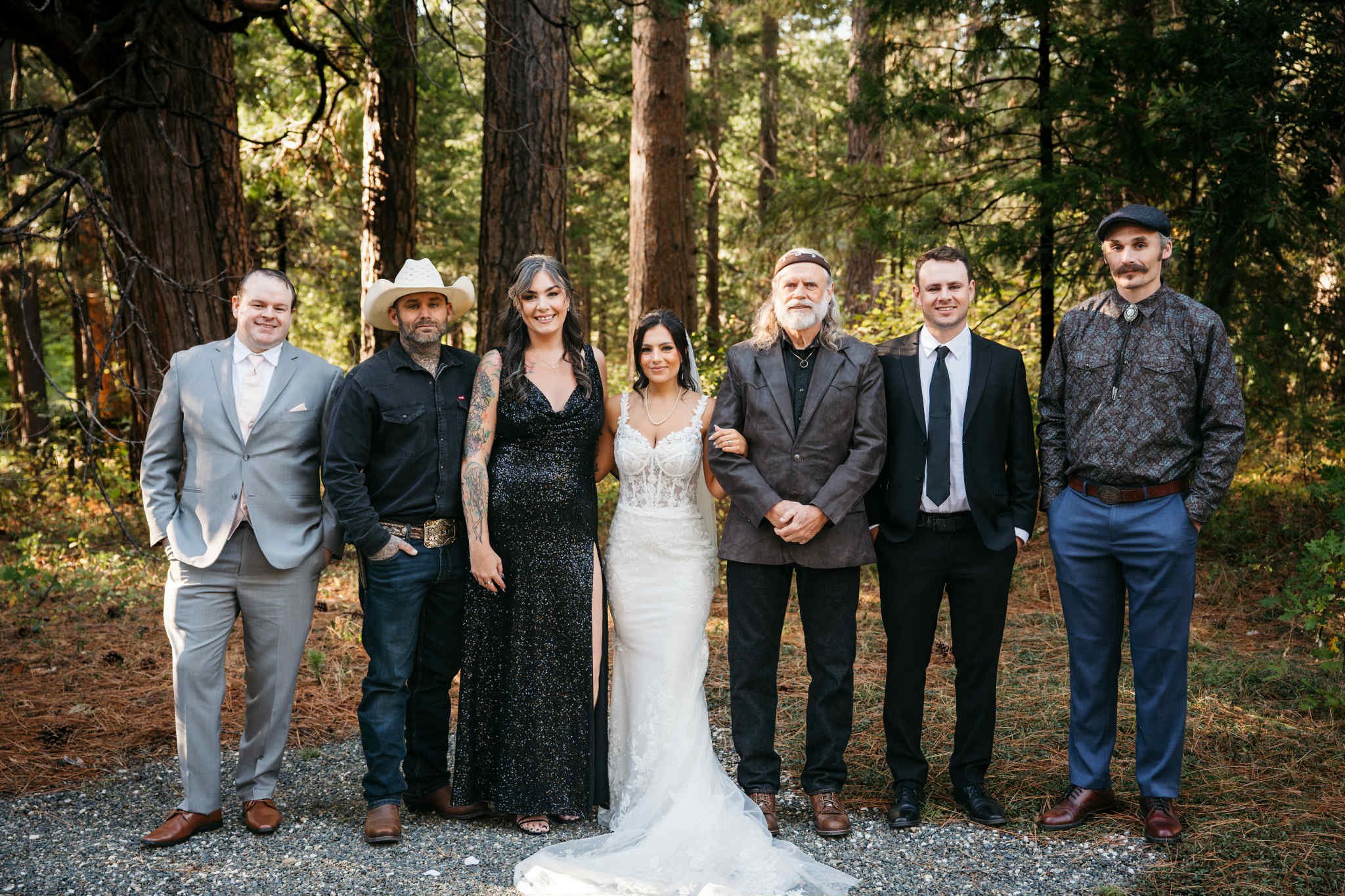 Group of seven people standing outdoors in a forest during daytime, dressed in formal and casual attire, including a bride in a white wedding gown, and a groom in a gray suit, posing for a photo.