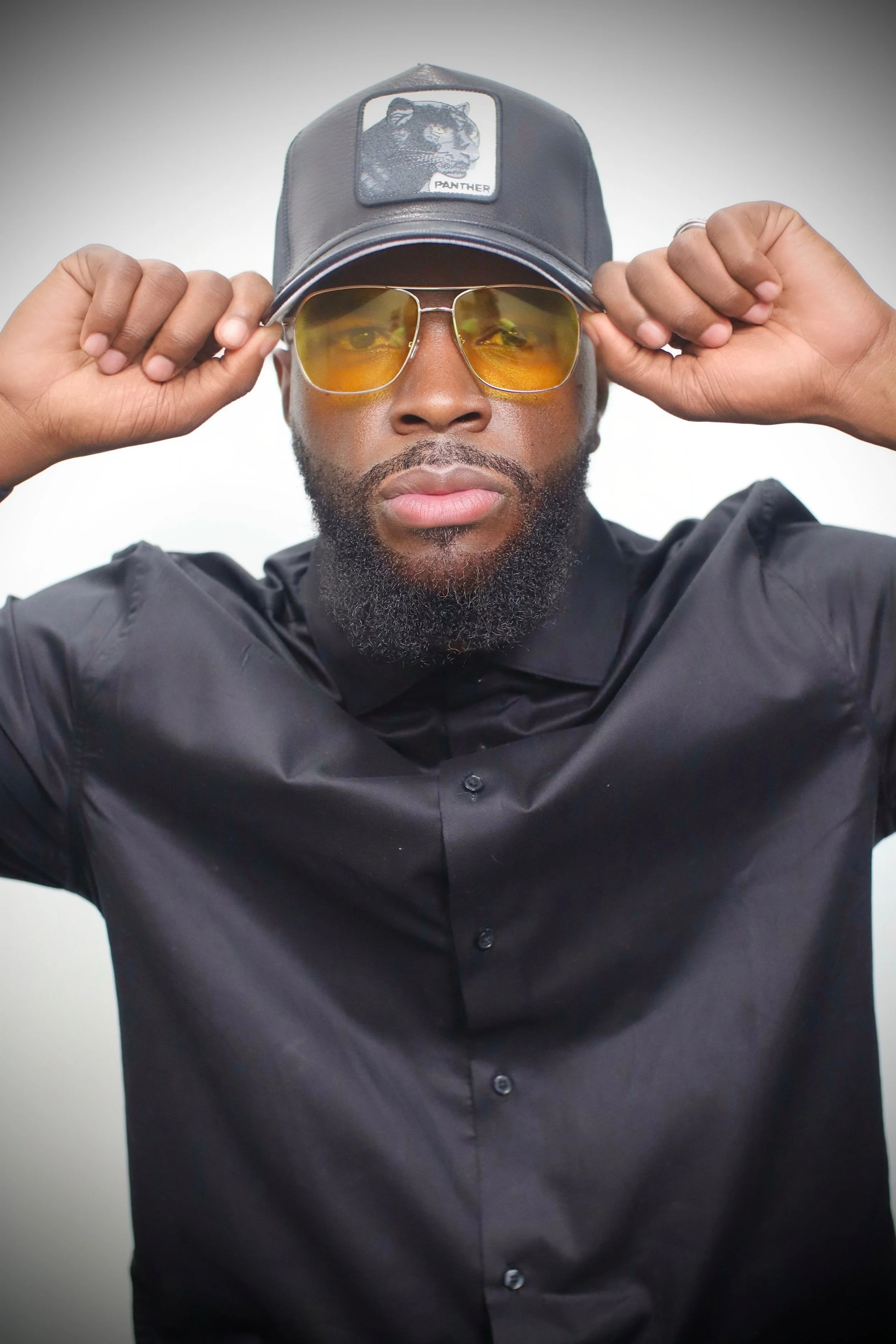 A man wearing a black leather cap with a panther patch, yellow tinted sunglasses, and a black button-up shirt.