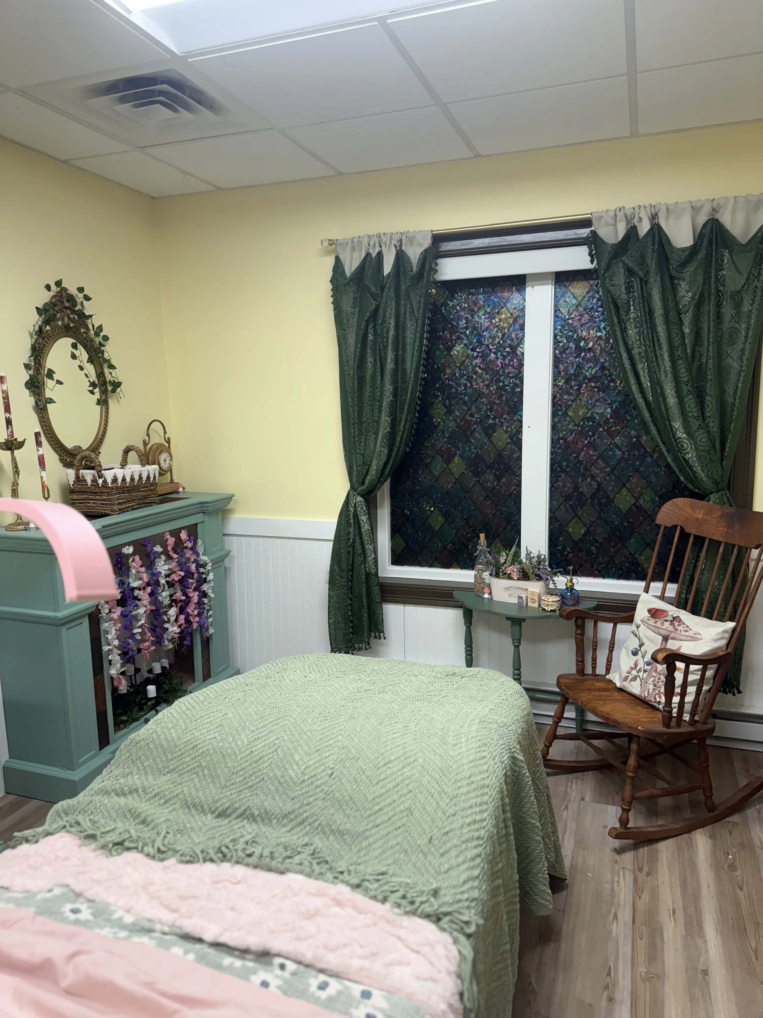 Cozy bedroom with a bed that has a green textured blanket, a blue dresser decorated with flowers and candles, a vintage mirror with foliage, a small green side table with bottles and flowers, dark green curtains with a colorful pattern, a wood rocking chair with a floral pillow, and wooden flooring.