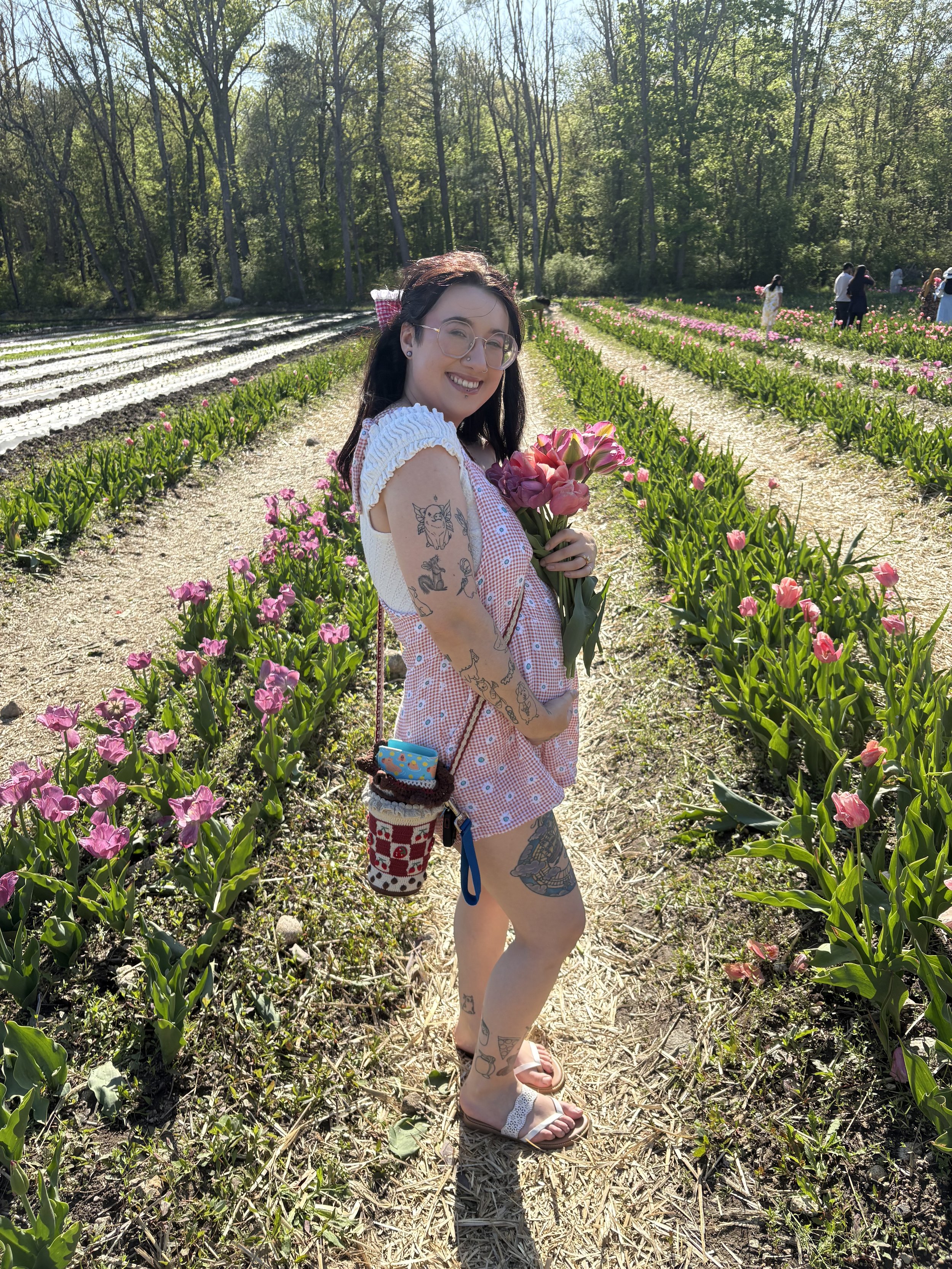 A smiling woman with glasses and tattoos, holding pink tulips, standing in a field of tulips on a sunny day with a group of people in the background.