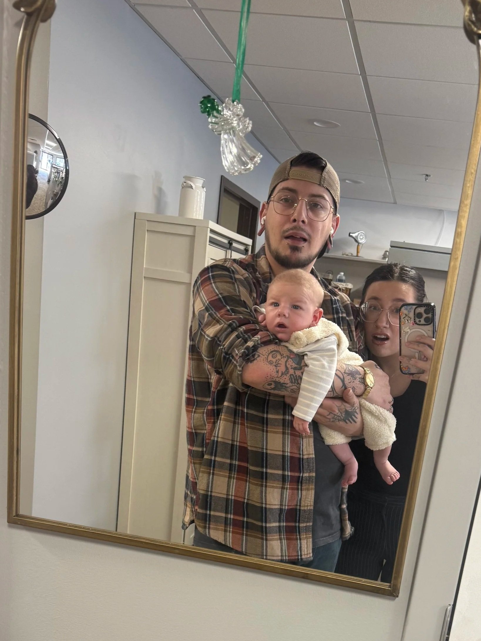 A person holding a baby taking a mirror selfie with another person partially visible behind him. The person holding the baby has tattoos on their arm, wears glasses, a baseball cap backward, and a plaid shirt. There is a balloon in the mirror reflection attached to the ceiling.