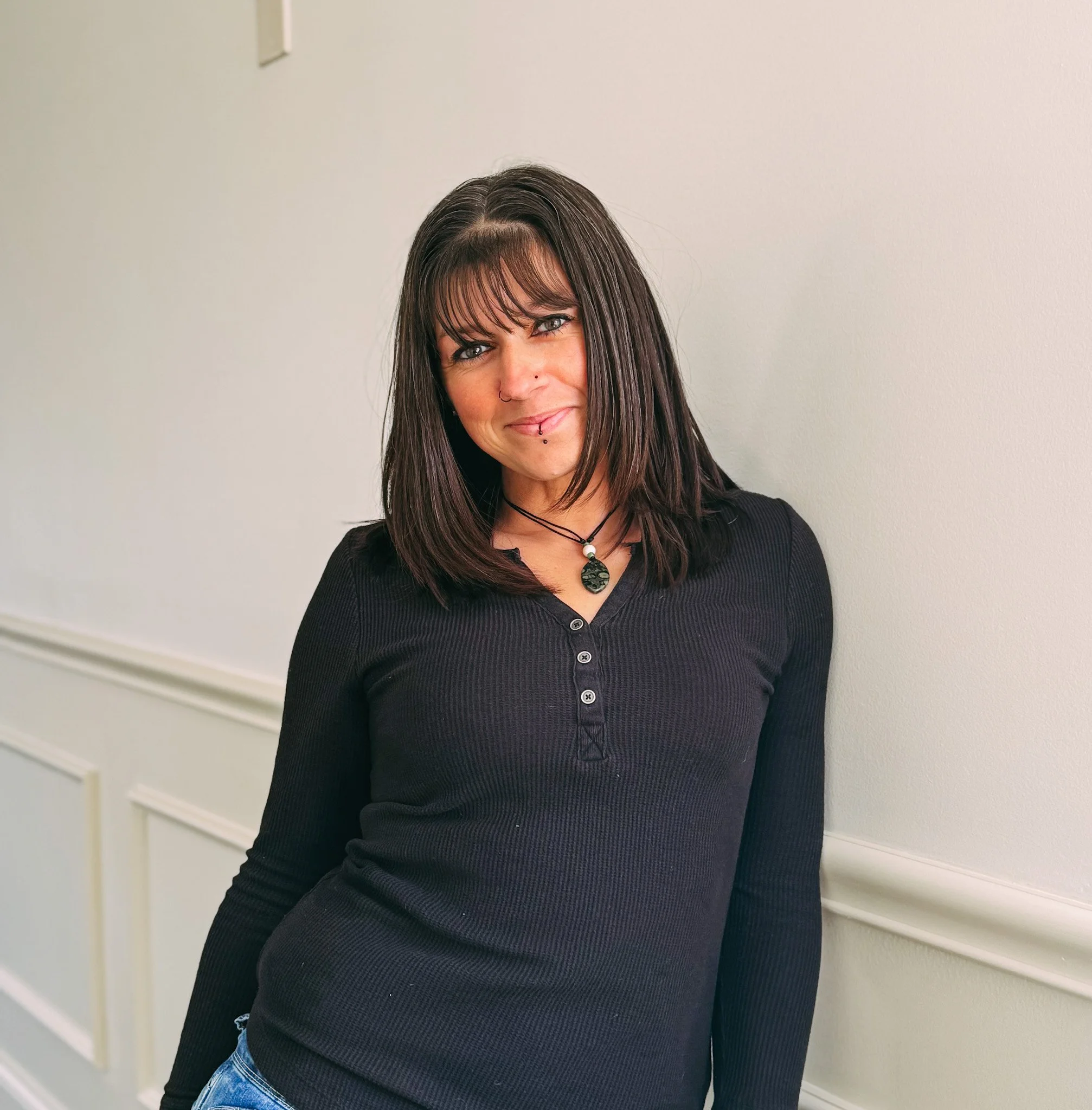 Woman with dark brown hair, wearing a black long sleeve top and blue jeans, standing against a plain wall.