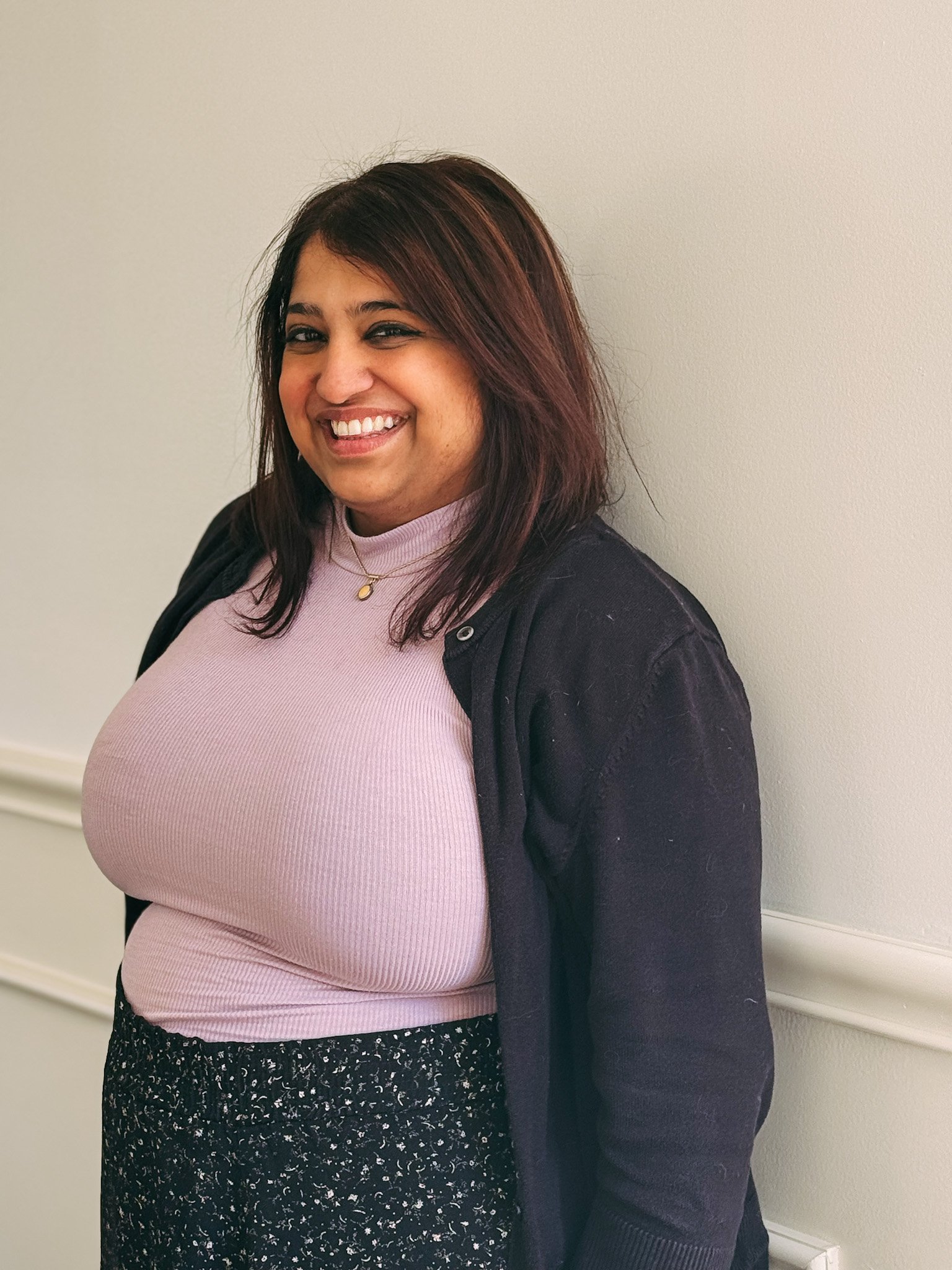 A woman with shoulder-length brown hair and light skin, smiling, wearing a pink ribbed turtleneck, black cardigan, and black patterned skirt, standing against a plain beige wall.