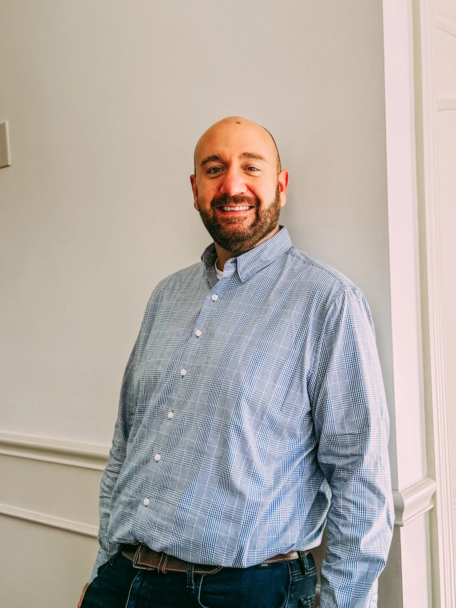 A smiling man with a beard and bald head wearing a checkered dress shirt and dark pants standing against a plain light-colored wall.