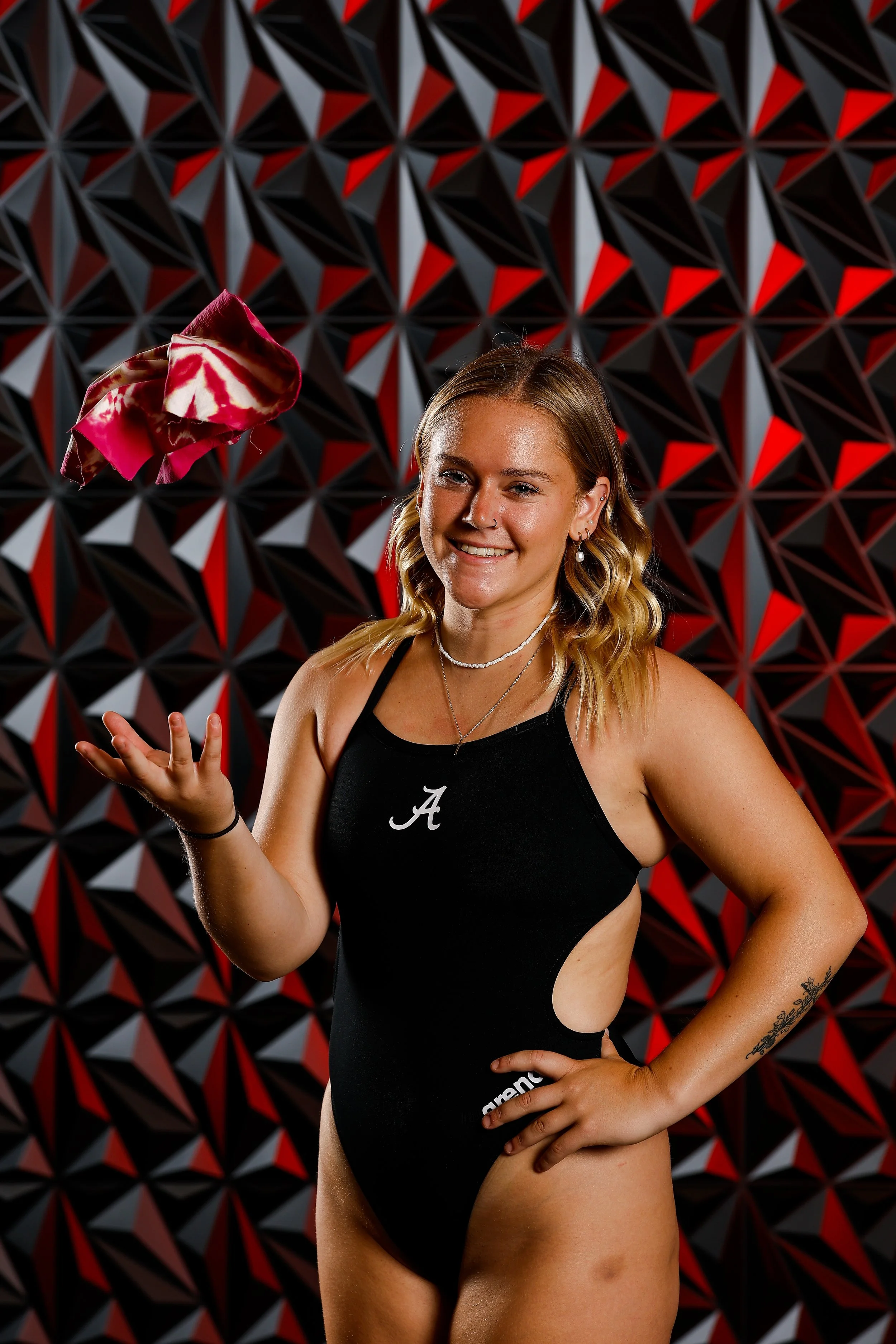 A young woman with blonde hair in waves, wearing a black athletic outfit with a white 'A' on the chest, smiling and standing in front of a red and black geometric patterned wall, tossing a red and white tie-dye bandana in the air.