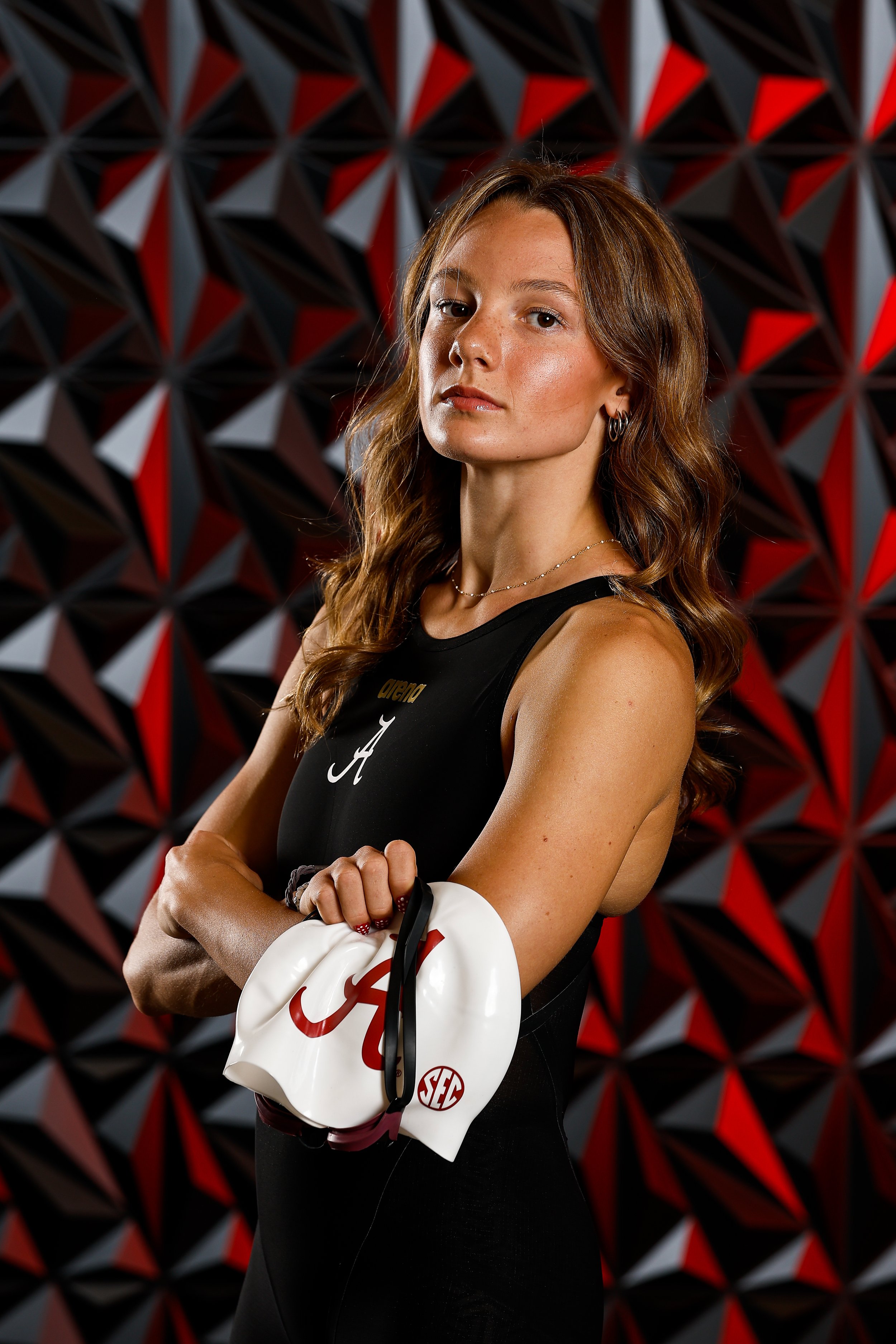 A female swimmer standing confidently against a geometric patterned wall, wearing a black athletic top, holding a white cap and a swim towel.