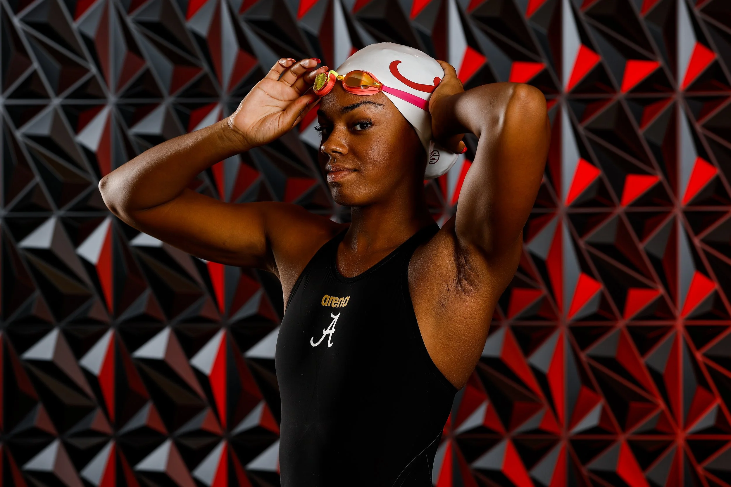 Female swimmer adjusting her swim cap and goggles, wearing a black arena swimsuit, standing in front of a geometric black, red, and gray background.