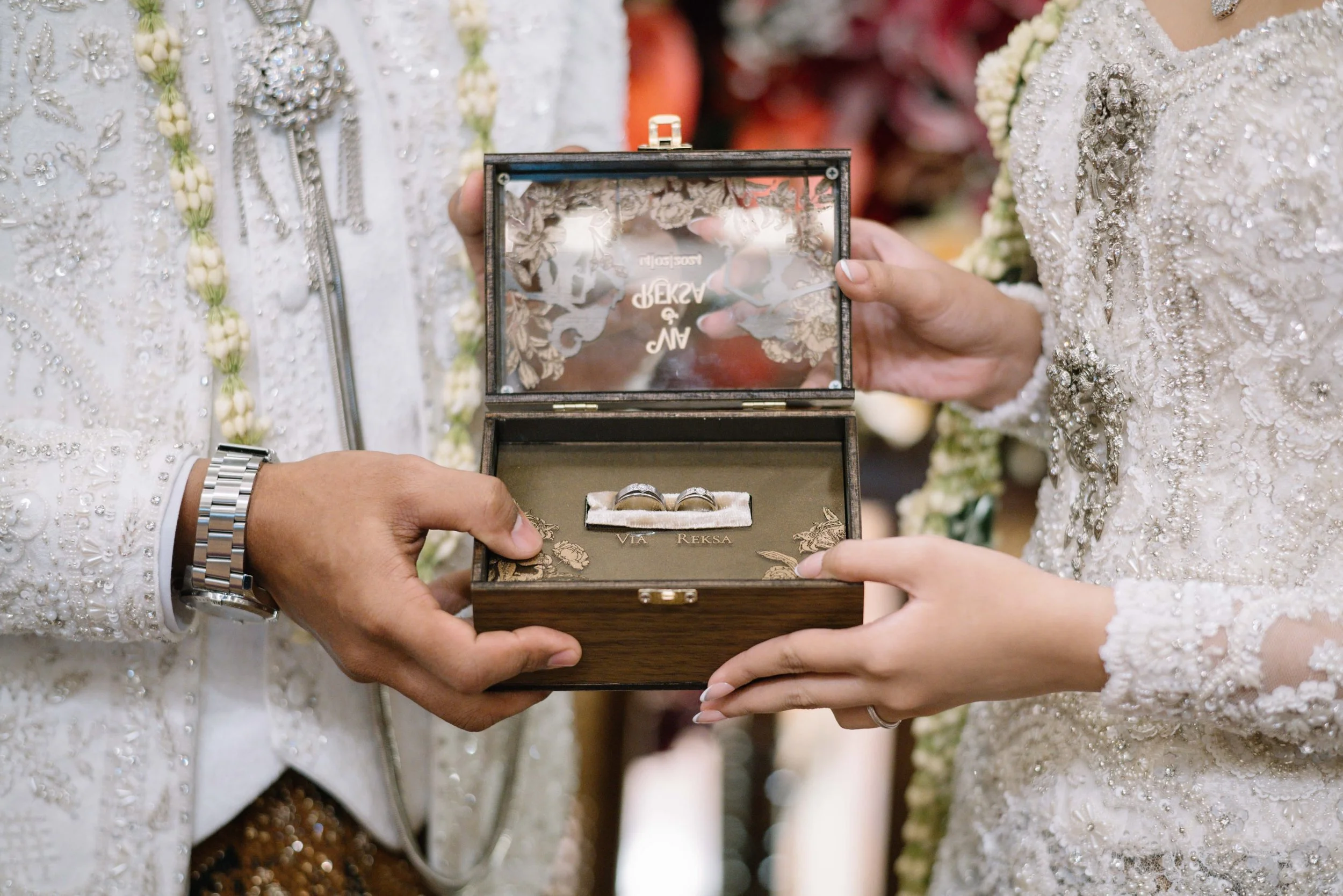 A person holding a wooden jewelry box with wedding rings inside during a wedding ceremony. Two people in traditional wedding attire are visible, both wearing ornate, embroidered white outfits with floral details and jewelry.