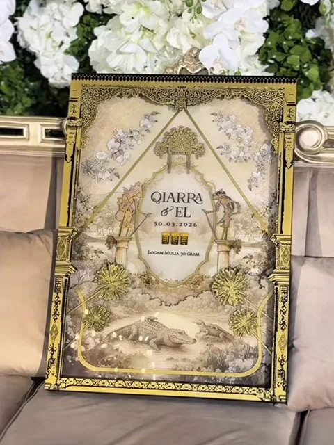 Invitation or sign with ornate gold and black border, floral design, date 10.03.2026, and names Qiarra and El, surrounded by white flowers and green foliage.