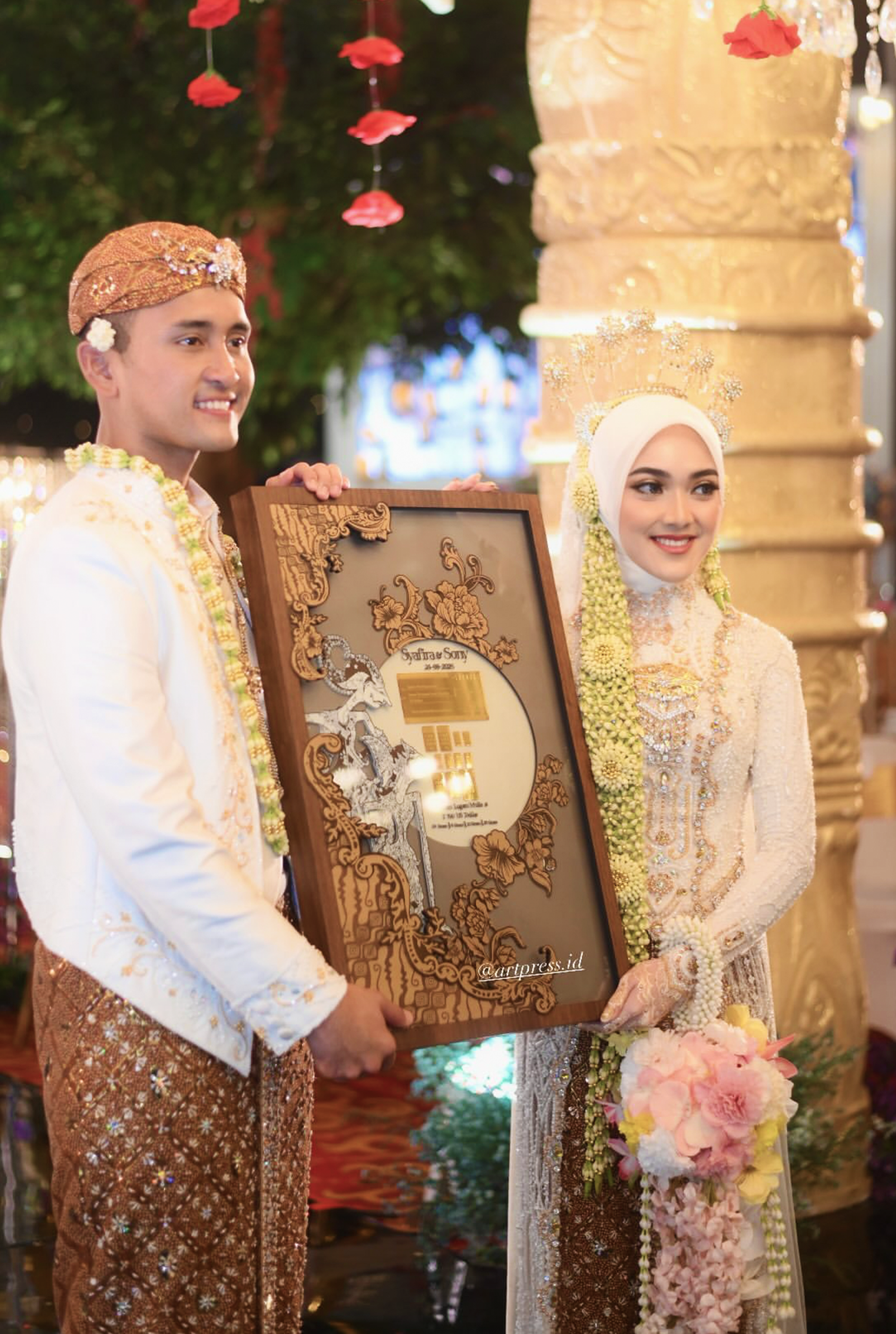 A couple in traditional Indonesian wedding attire holding a framed certificate or artwork. The groom is wearing a white and gold outfit with a brown and gold headpiece. The bride wears a white dress with intricate gold embellishments, a white hijab, and floral garlands. They are smiling during a wedding ceremony decorated with hanging red flower petals, floral arrangements, and warm lighting.