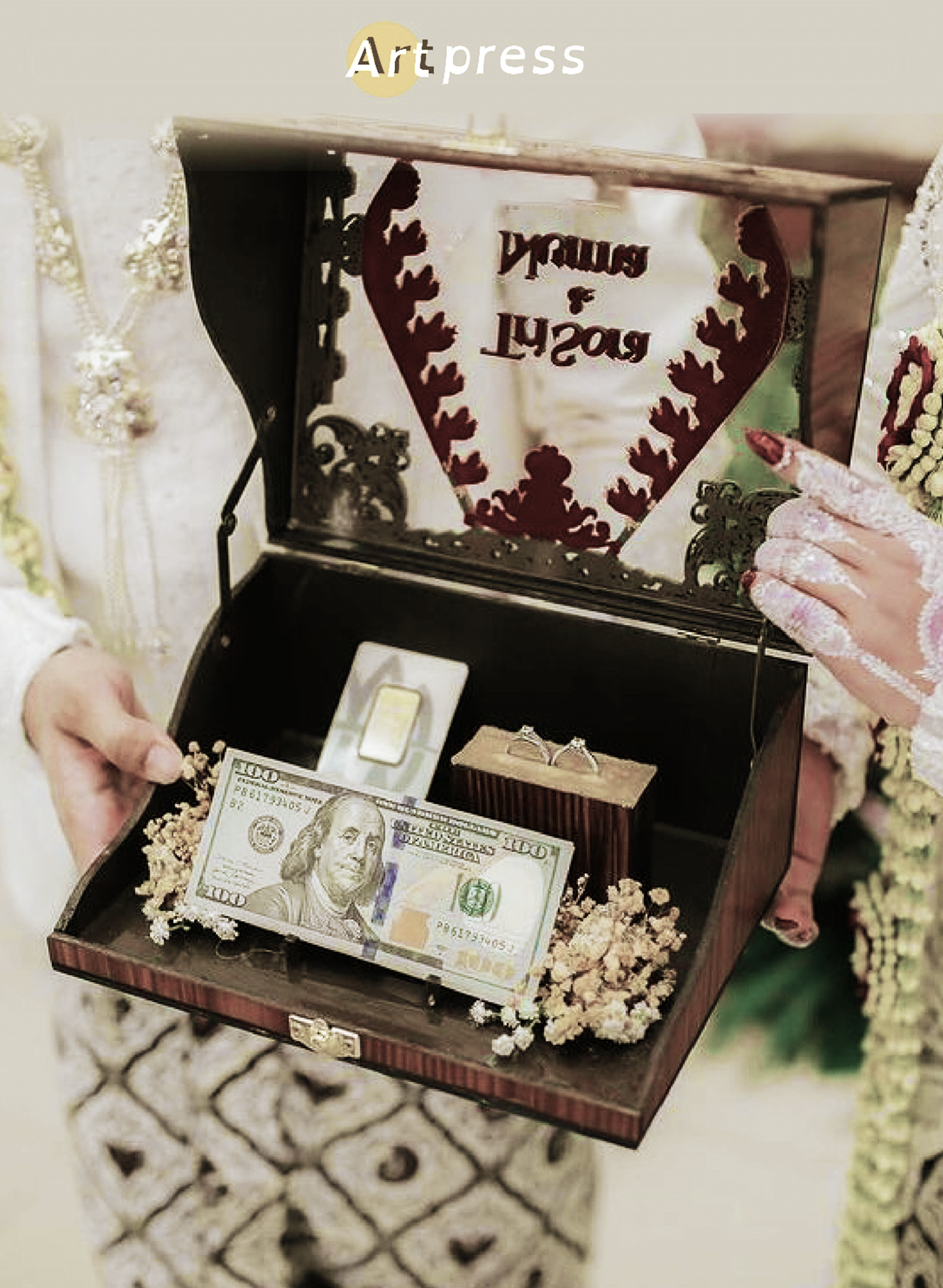 A jewelry box with a hundred-dollar bill, decorated with white flowers, and a person's hand holding the box, with a background of grass and a patterned surface.