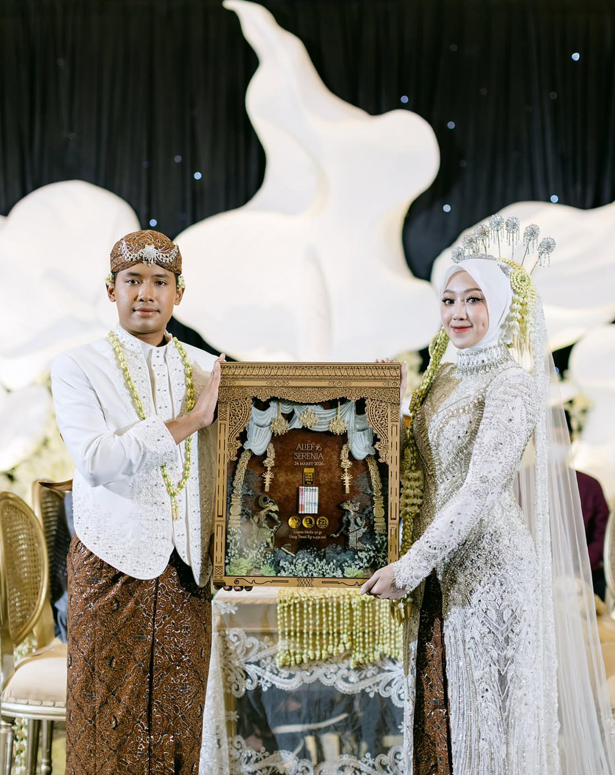 A bride and groom in traditional Indonesian wedding attire holding a framed wedding certificate at their wedding reception.