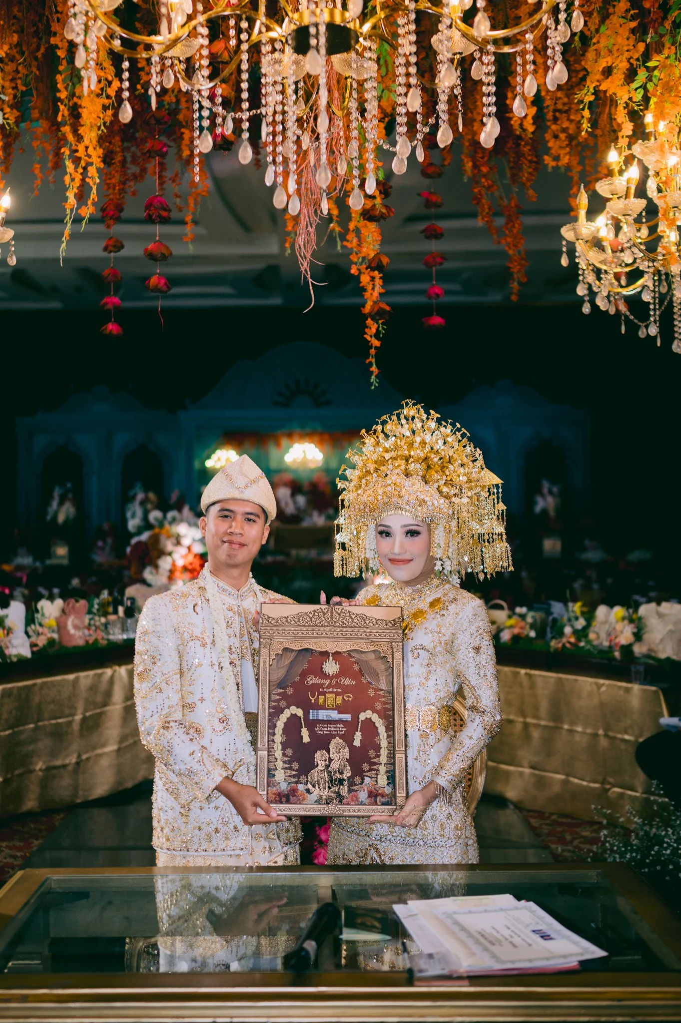 A traditional wedding ceremony with a bride and groom dressed in ornate white and gold attire, holding a framed certificate, standing under a decorated ceiling with hanging flowers and chandeliers, in a beautifully decorated venue.