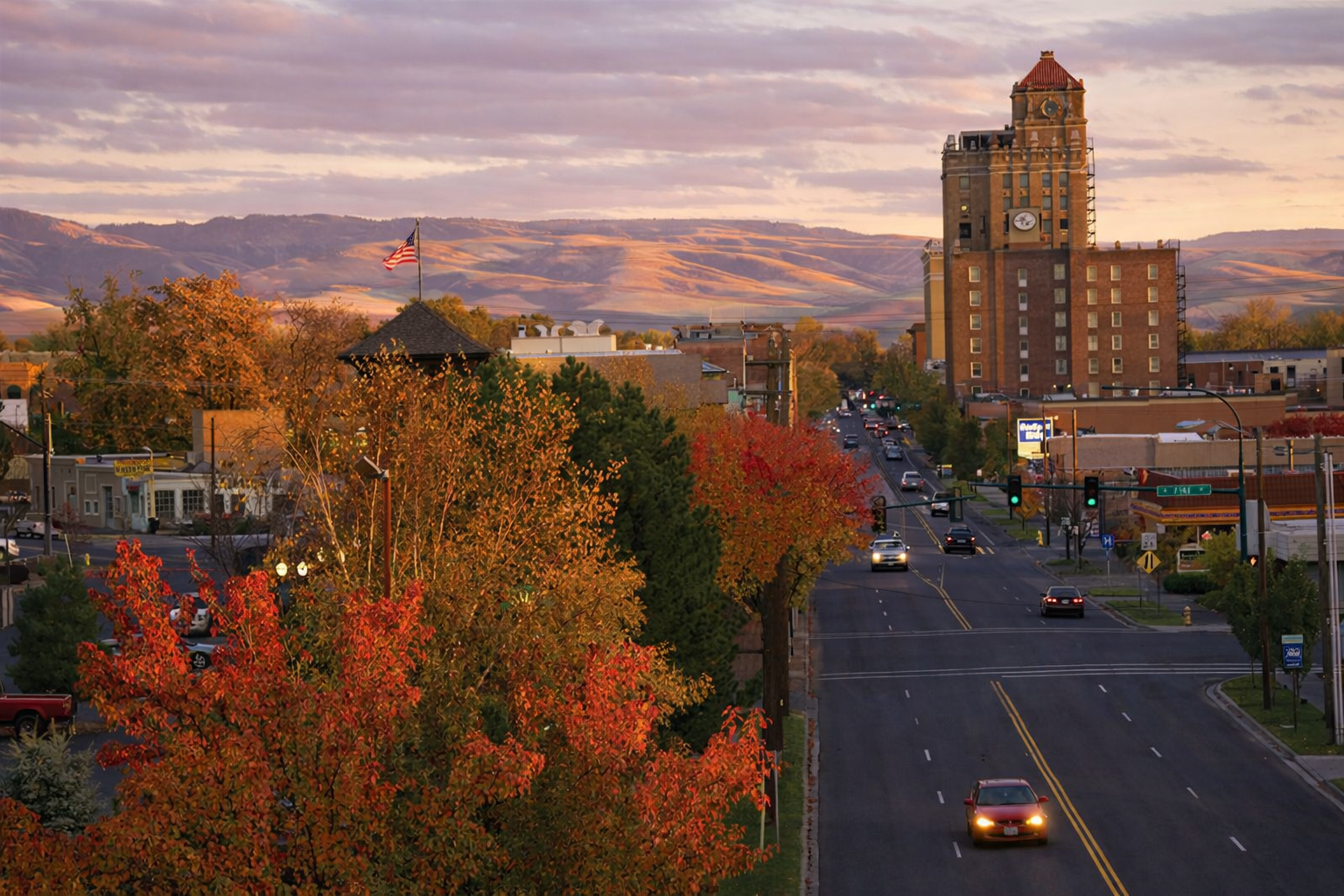 A small city street during sunset with cars driving on the road. There are colorfully autumn trees lining the street, a clock tower in the background, and rolling hills with a sunset sky.