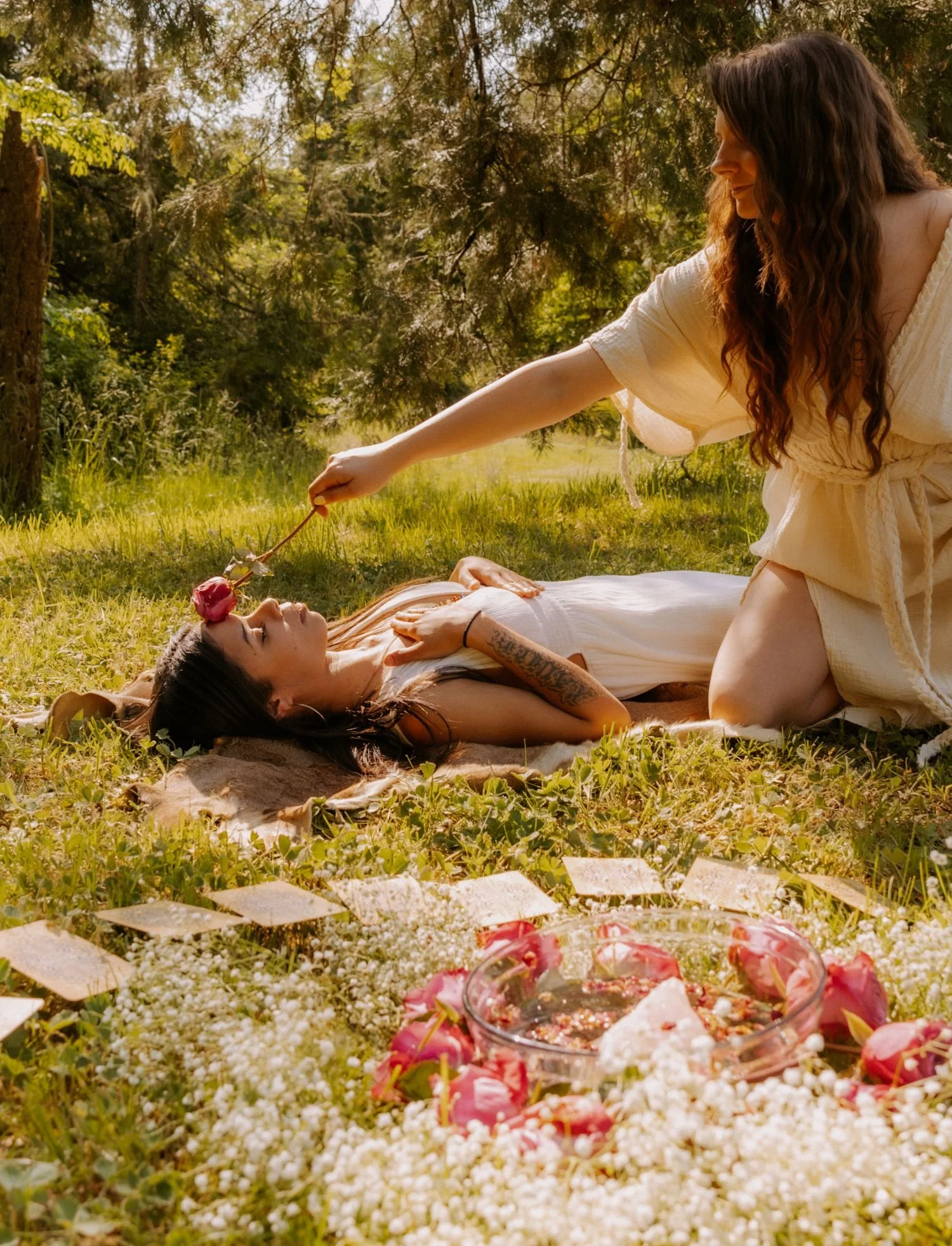 A woman in a white dress laying on the grass with her eyes closed, while another woman kneels next to her, holding a rose over her face in an outdoor setting surrounded by trees and flowers. There is a floral arrangement with a glass bowl of water an