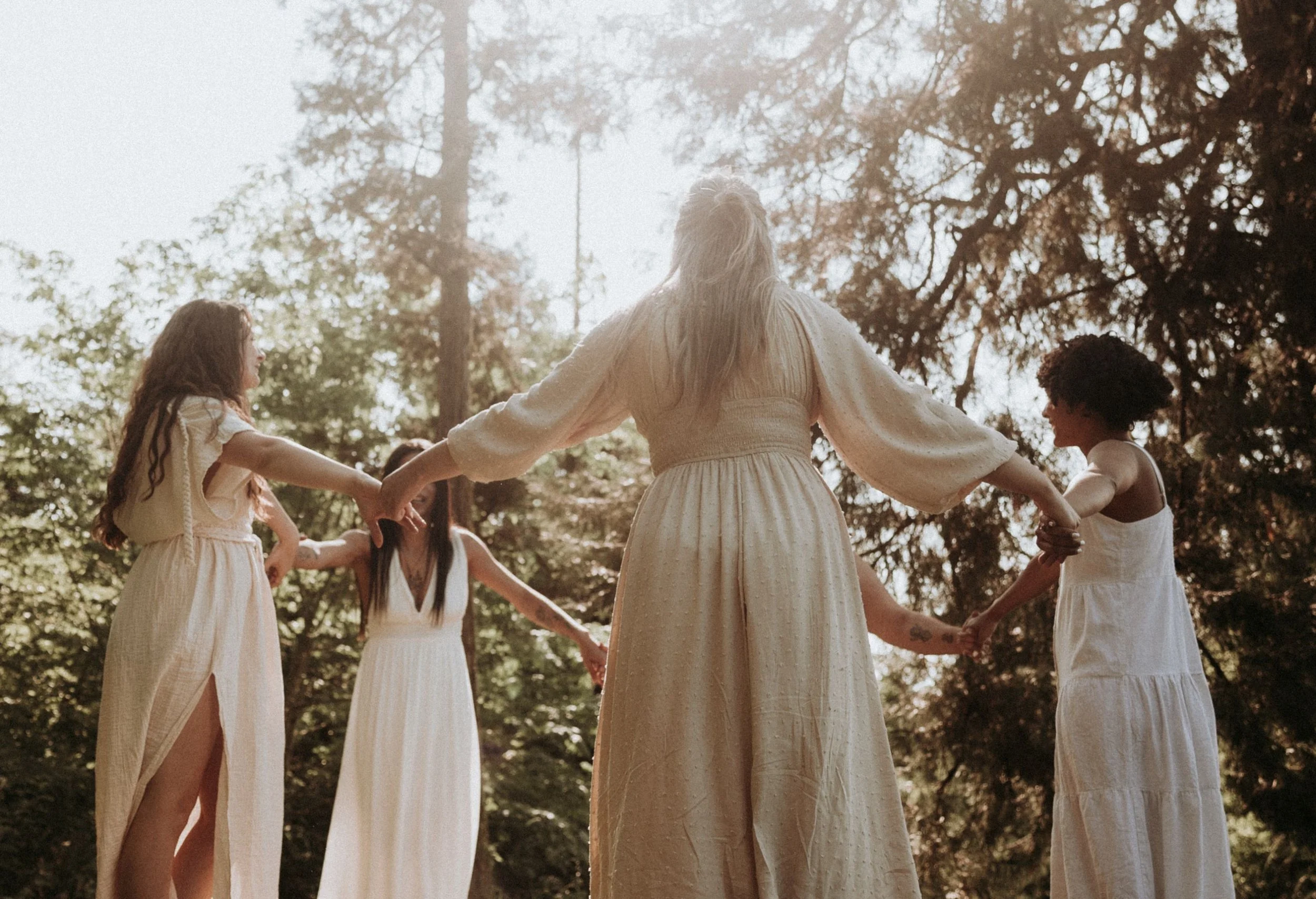 A group of women holding hands in a circle outdoors surrounded by trees during daylight.