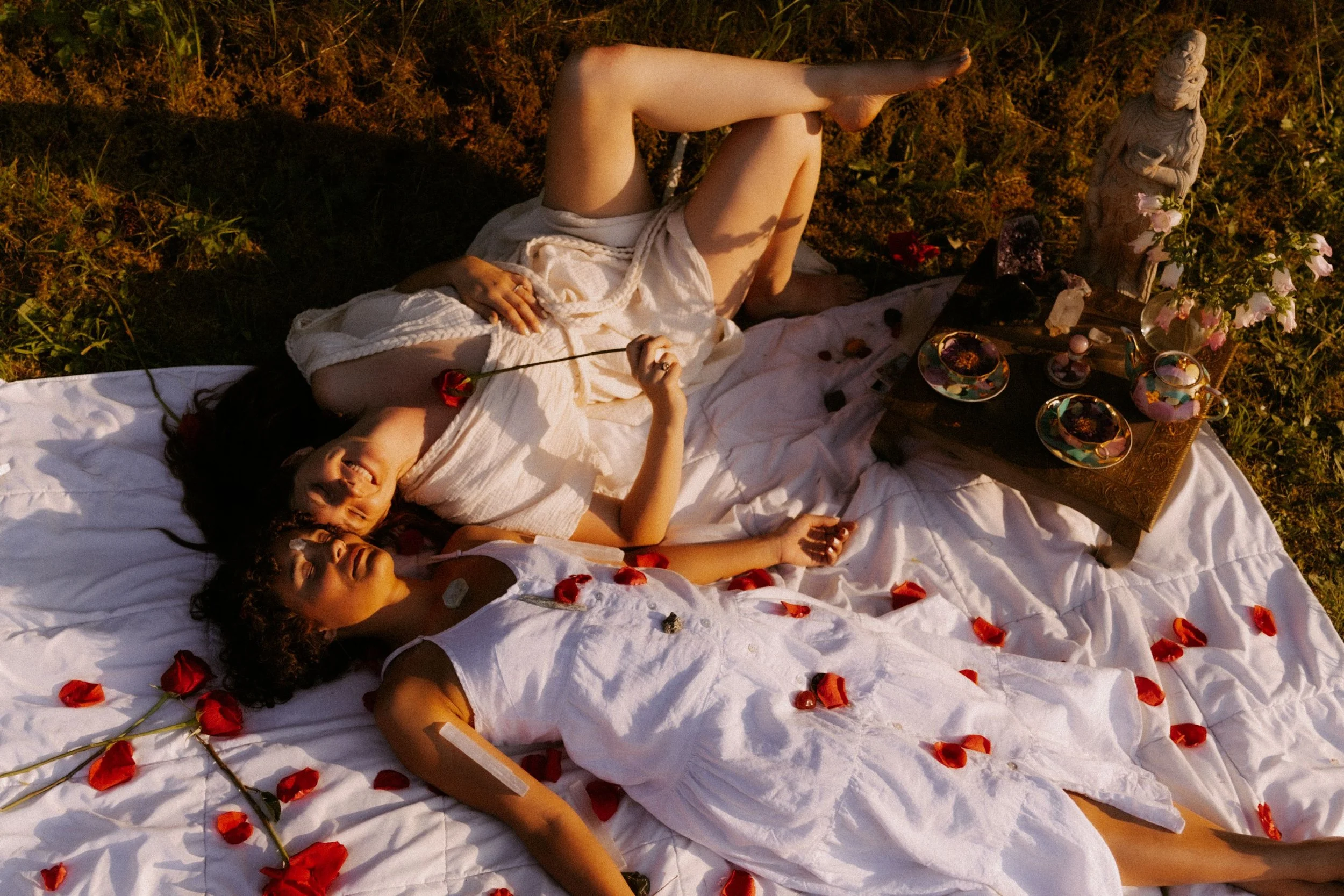 Two women in white dresses lying on a white sheet outdoors surrounded by rose petals, with a small altar decorated with flowers and statues nearby.