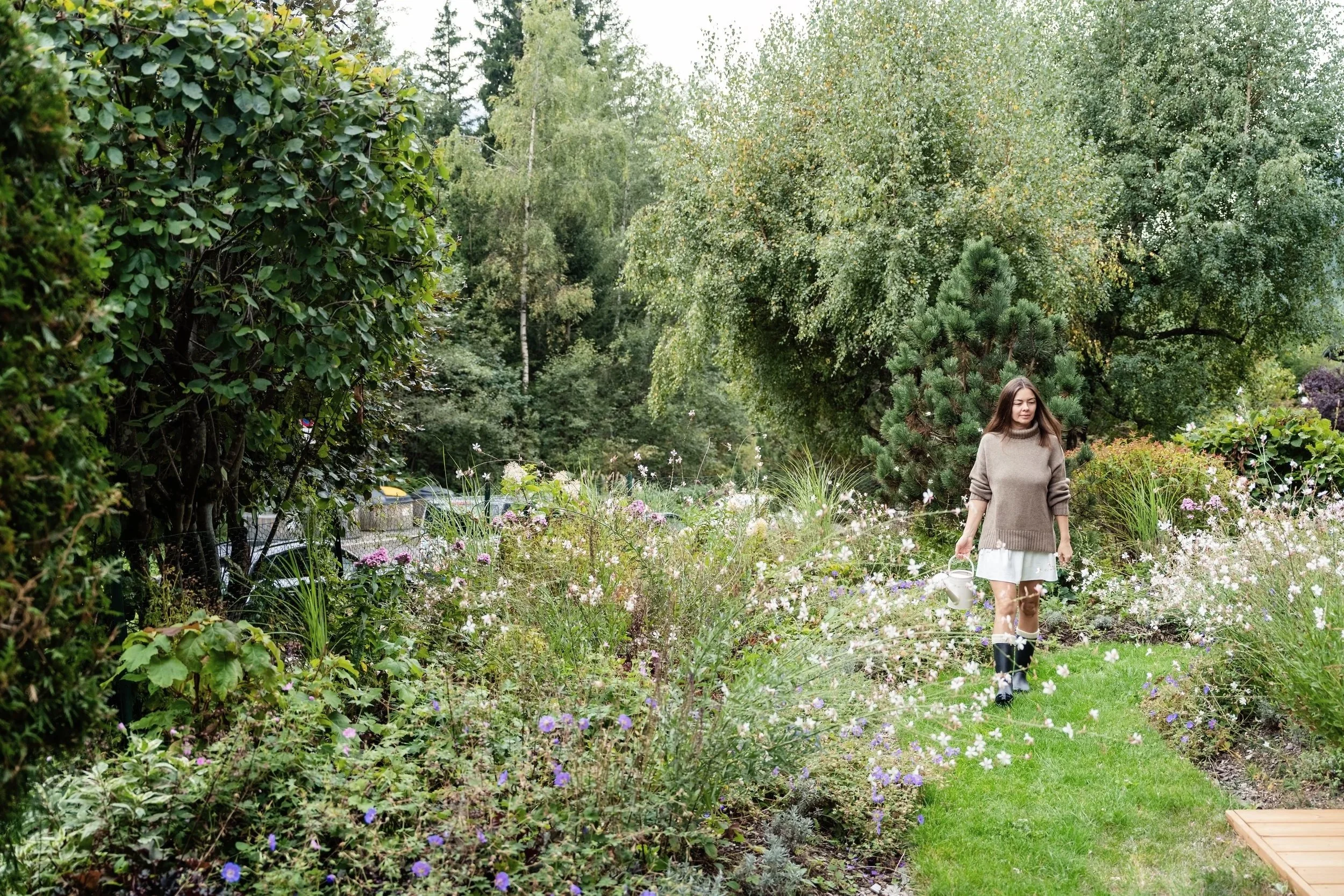A woman walking on a grassy path in a garden with various green trees and colorful flowers, holding a watering can.
