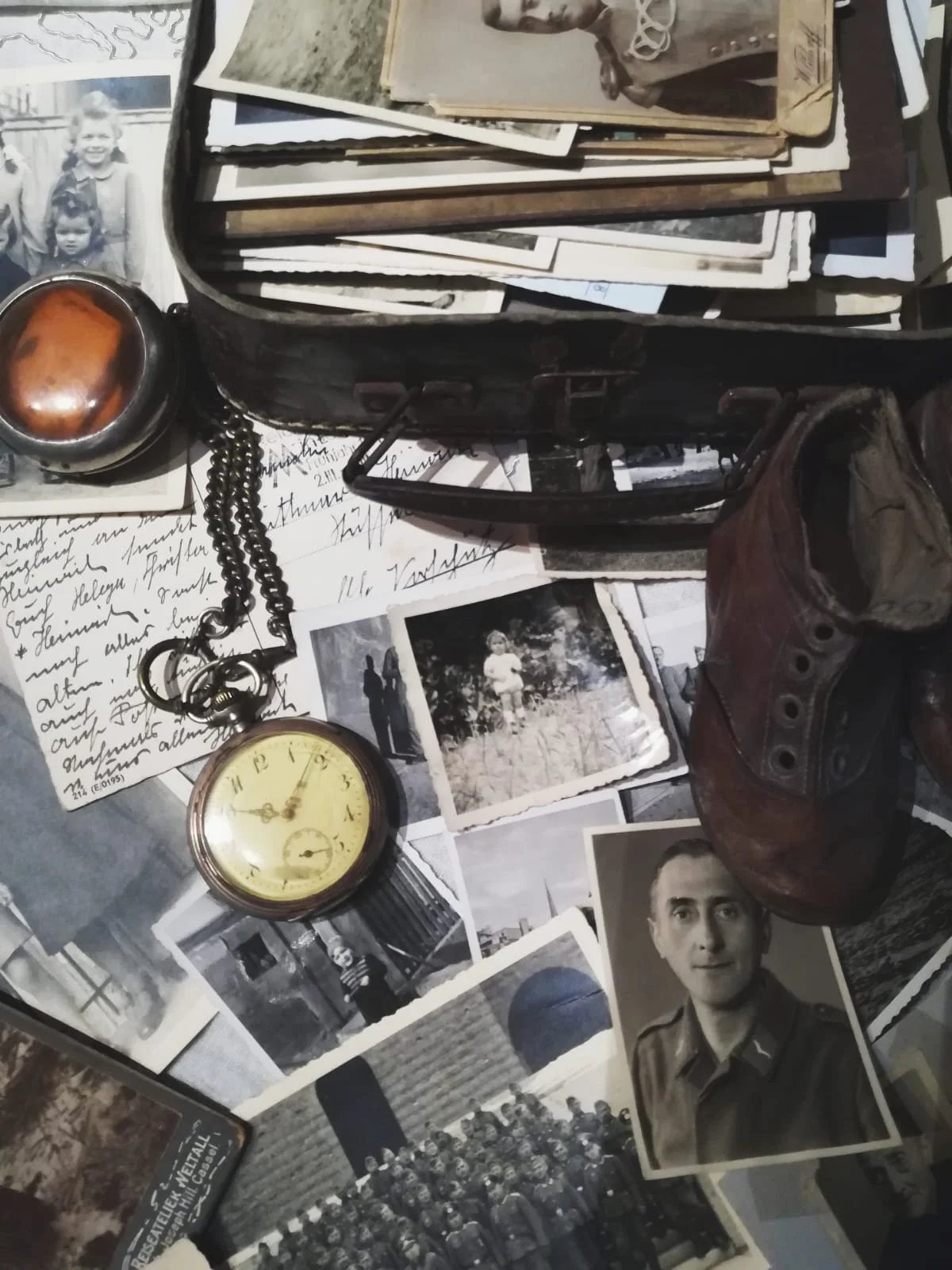 Alte Schwarz-Weiß-Fotografien, eine Taschenuhr, ein alter Stiefel, ein Zigarillokästchen und ein Kettenanhänger sind auf einer Tischoberfläche verteilt.