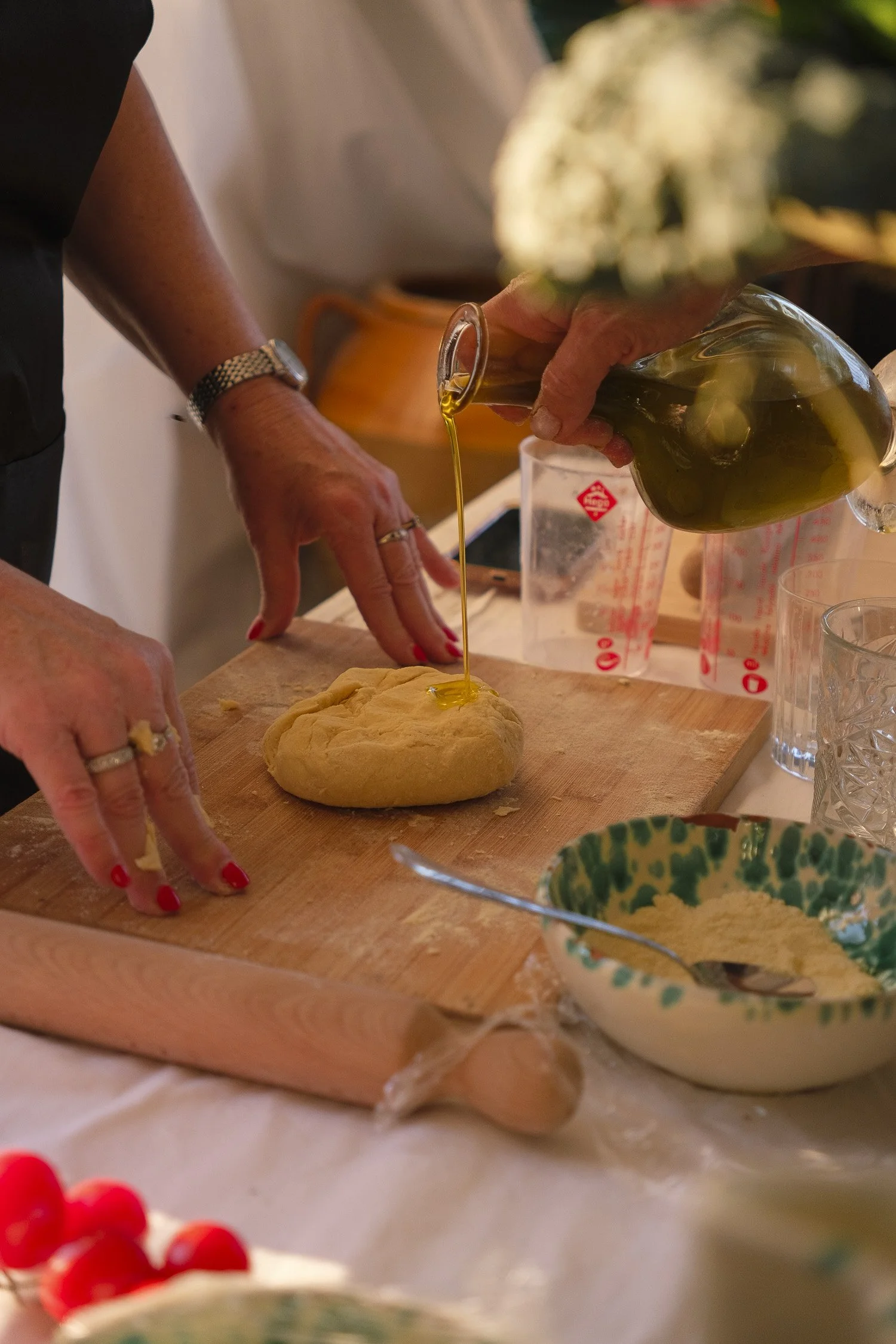 Person pouring olive oil onto dough on a wooden board during food preparation.